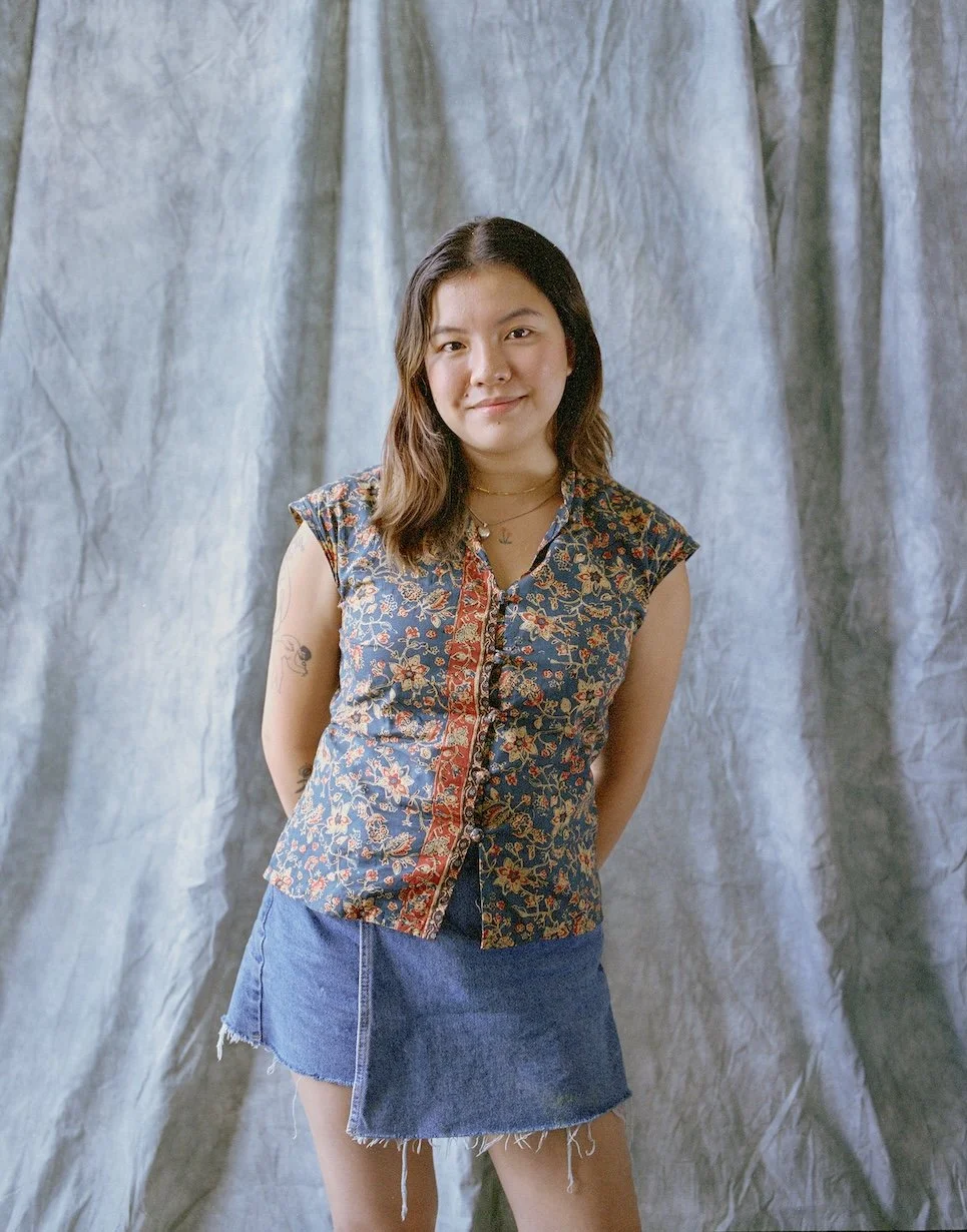 A young woman with shoulder-length dark hair, wearing a floral patterned sleeveless shirt and a denim skirt, standing in front of a gray fabric backdrop.