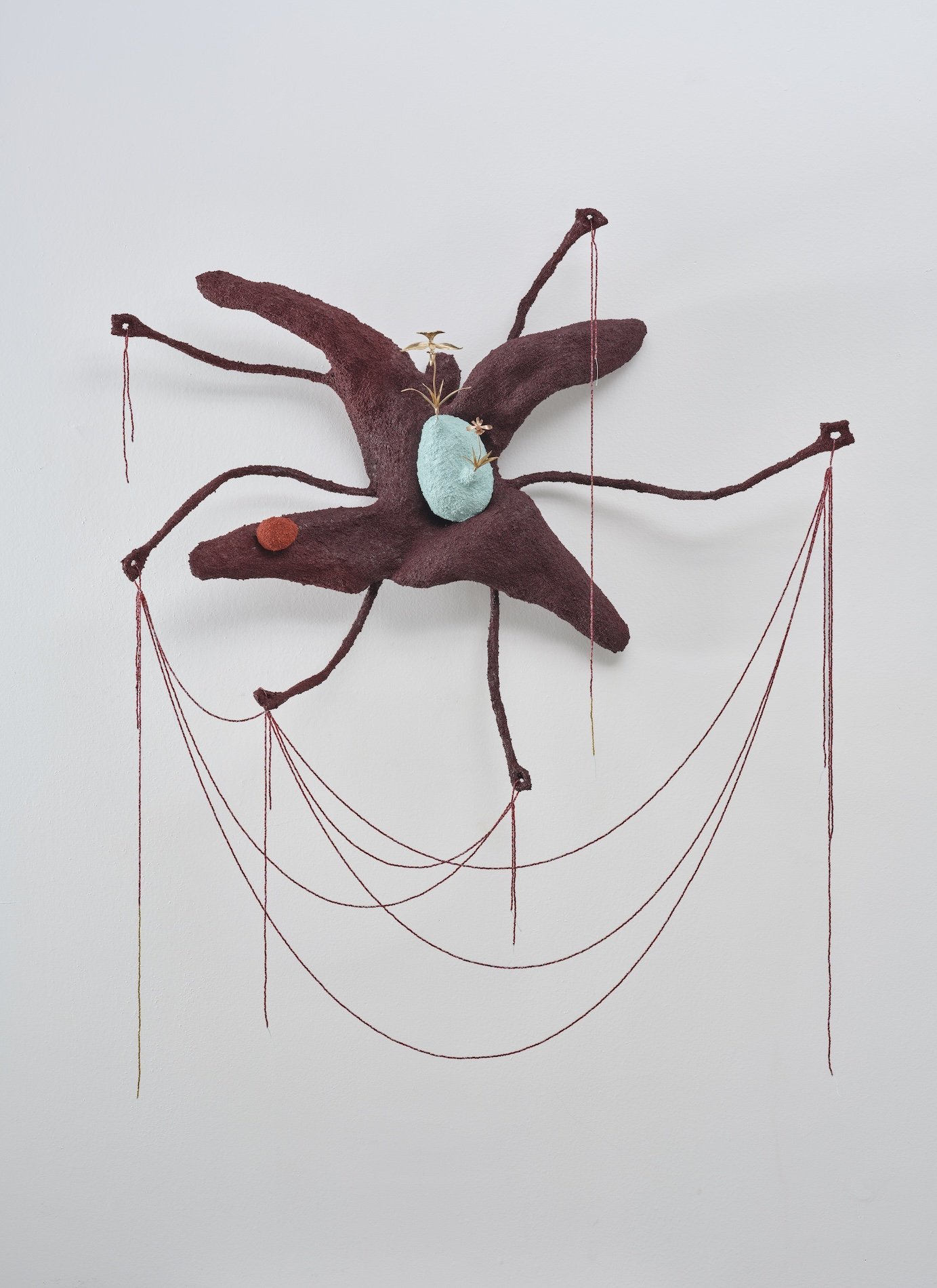 Mixed media art piece featuring a purple star-shaped form with extended arms, a turquoise oval shape at the center, gold and pink accents, and red strings hanging from the arms, all mounted on a white wall.