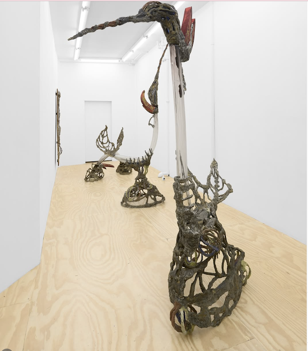 Sculpture resembling a tall, elongated figure with an abstract, twisted design, displayed in an art gallery with white walls and wooden floor.
