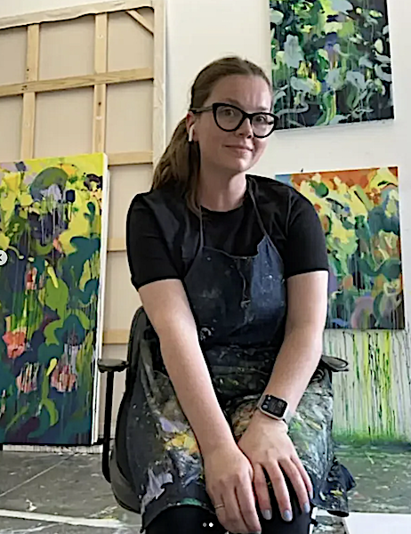 A woman with glasses and a ponytail sitting in front of colorful abstract paintings in an art studio.