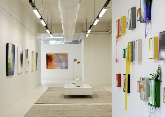Art gallery featuring colorful abstract paintings and mixed media on white walls, with a central seating area and modern lighting.