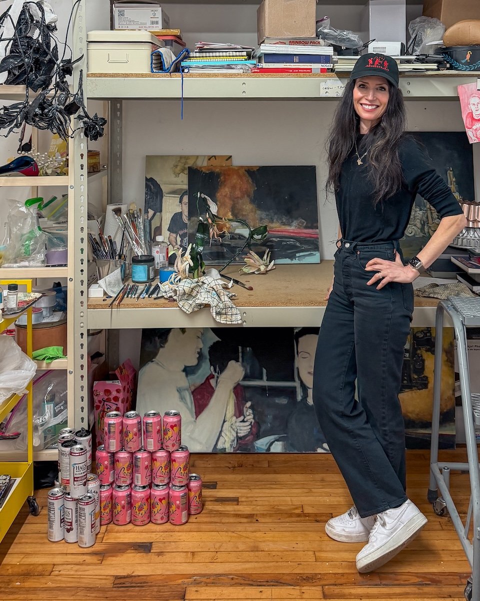 A woman wearing a black cap, black shirt, black jeans, and white sneakers stands in an art studio or workspace, smiling with her hands on her hips. Behind her are paintings, art supplies, and shelves with various items, including a stack of pink cans on the wooden floor.