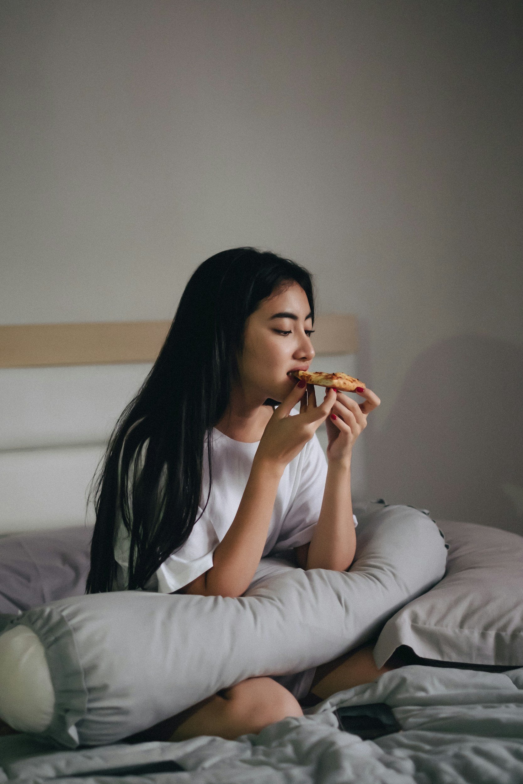 A female teenager eating a piece of pizza while sitting cross-legged on a bed