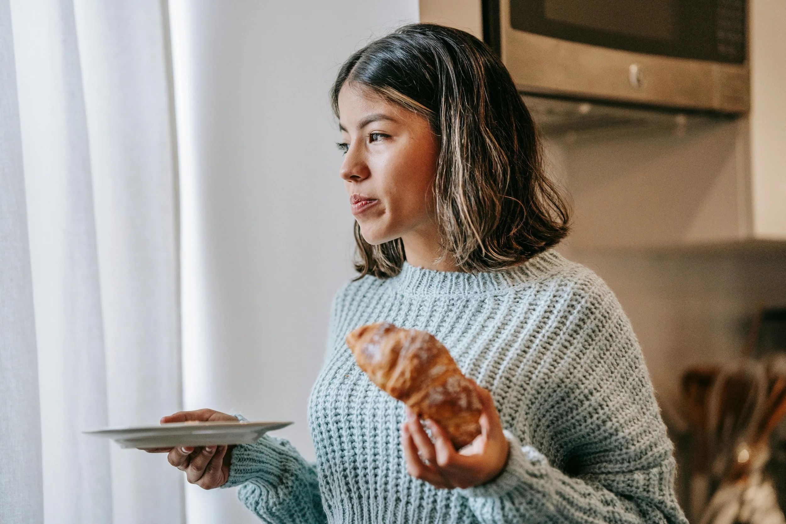 A woman holding a plate and a croissant while looking out the window