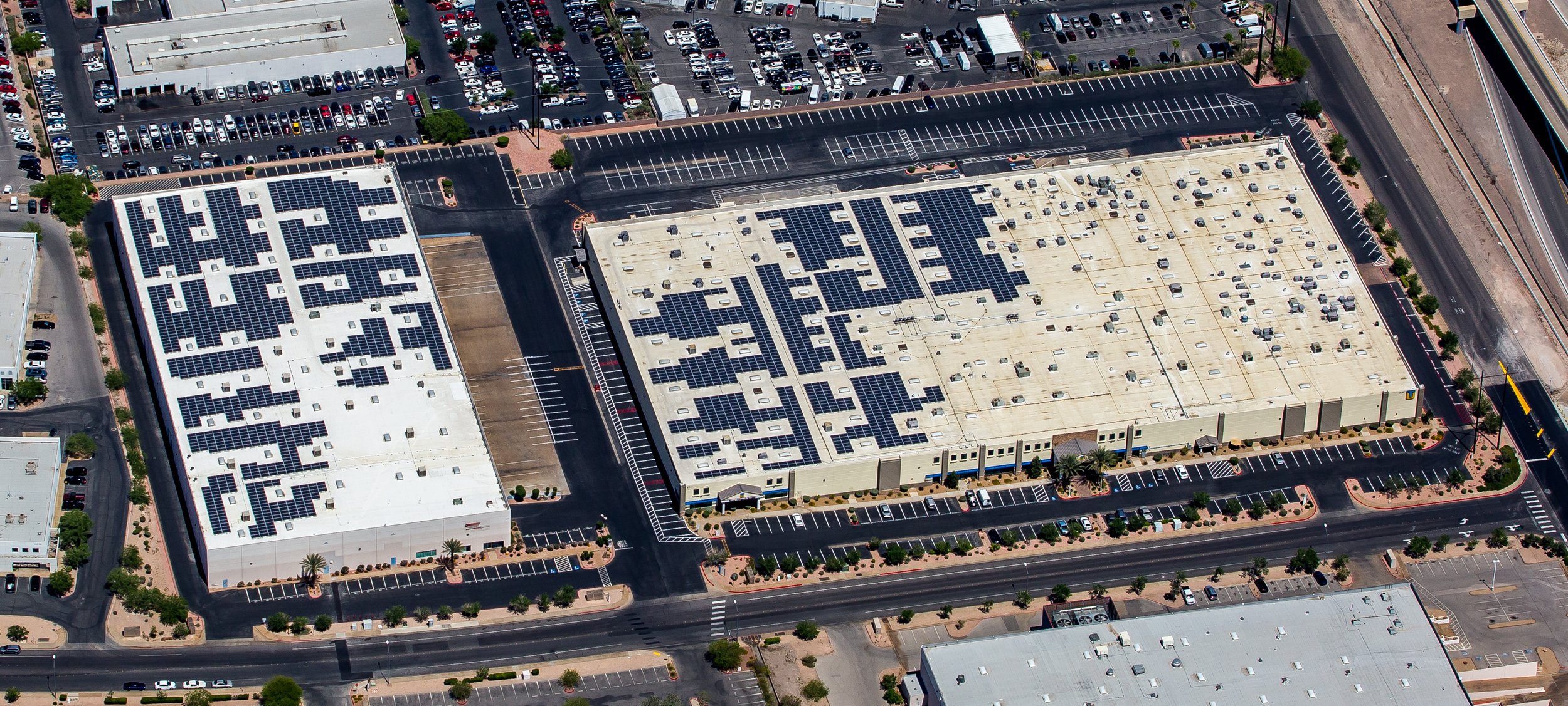 An aerial view of a large commercial building with numerous solar panels on the flat rooftops, surrounded by parking lots, streets, and some landscaped areas with trees.