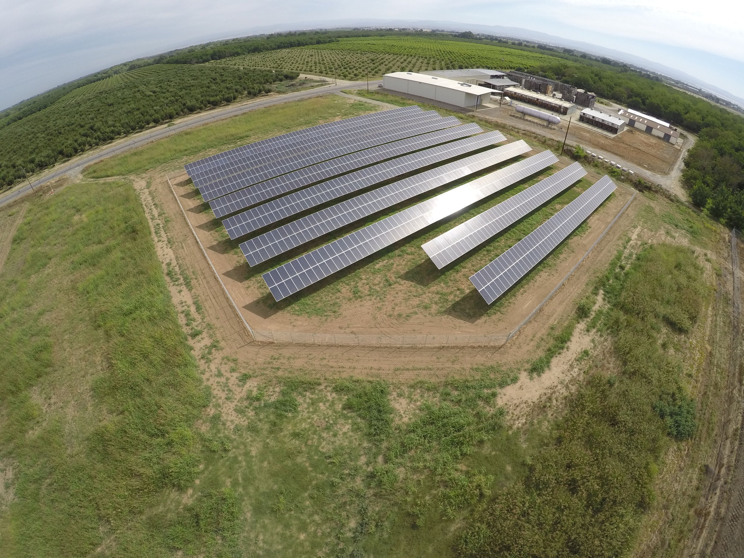 Aerial view of a solar farm with multiple rows of solar panels surrounded by green fields and trees, with industrial buildings in the background.