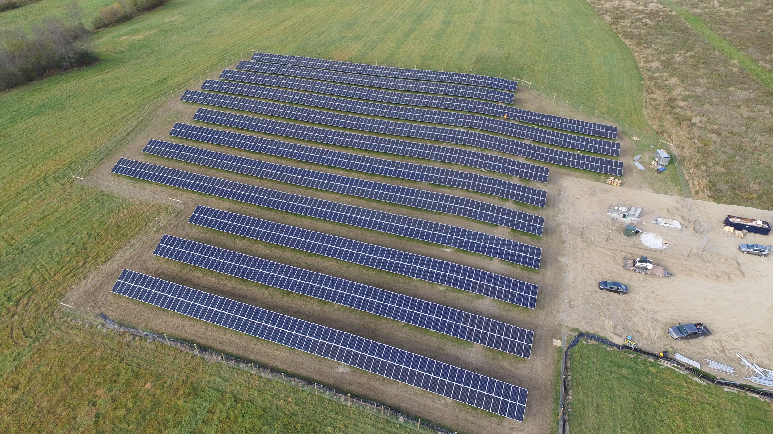 Aerial view of a solar farm with multiple rows of solar panels installed on a grassy field. There are a few vehicles and equipment near the edge of the installation.