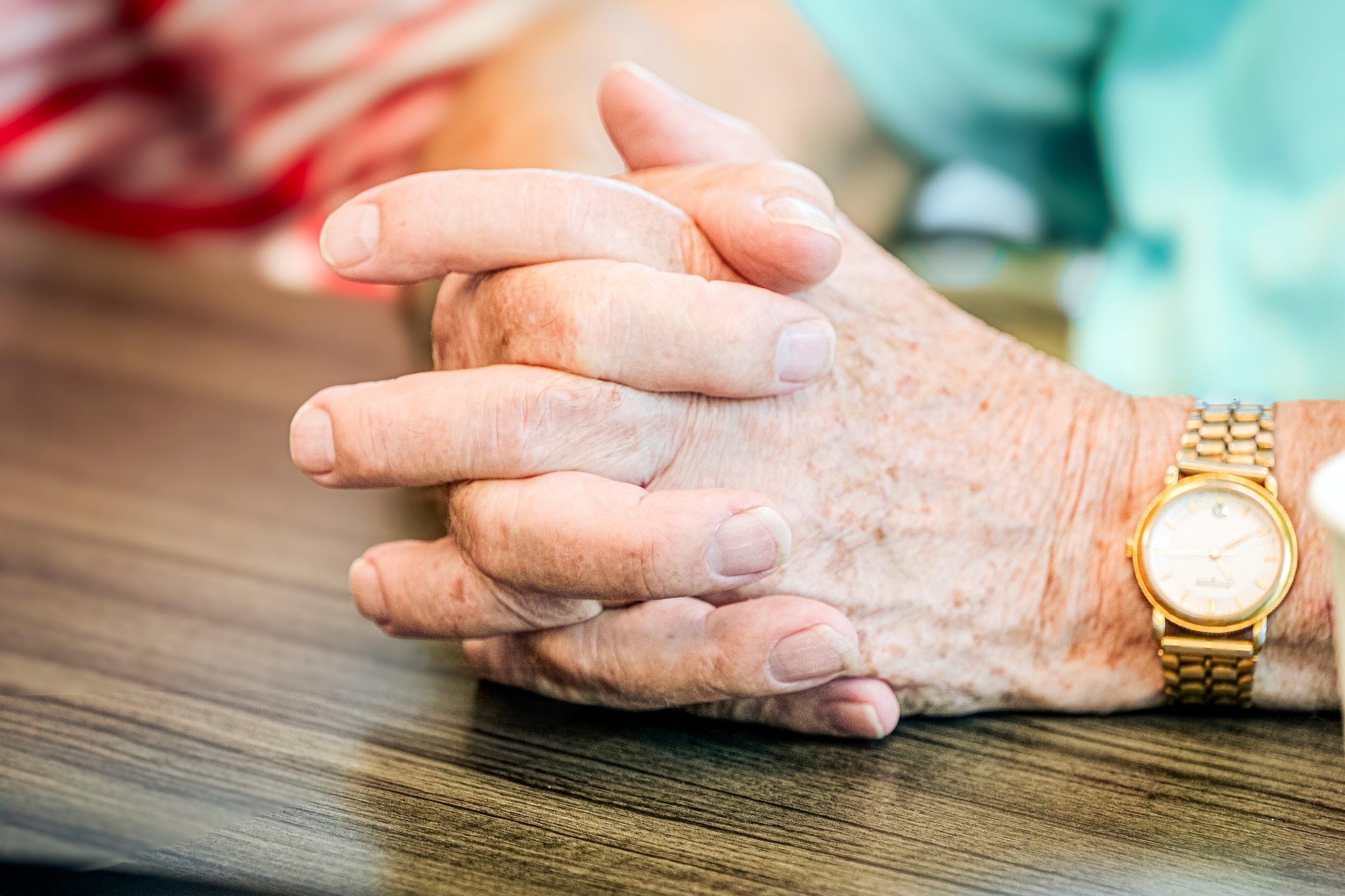 Close-up of clasped hands with a gold wristwatch, resting on a wooden table.