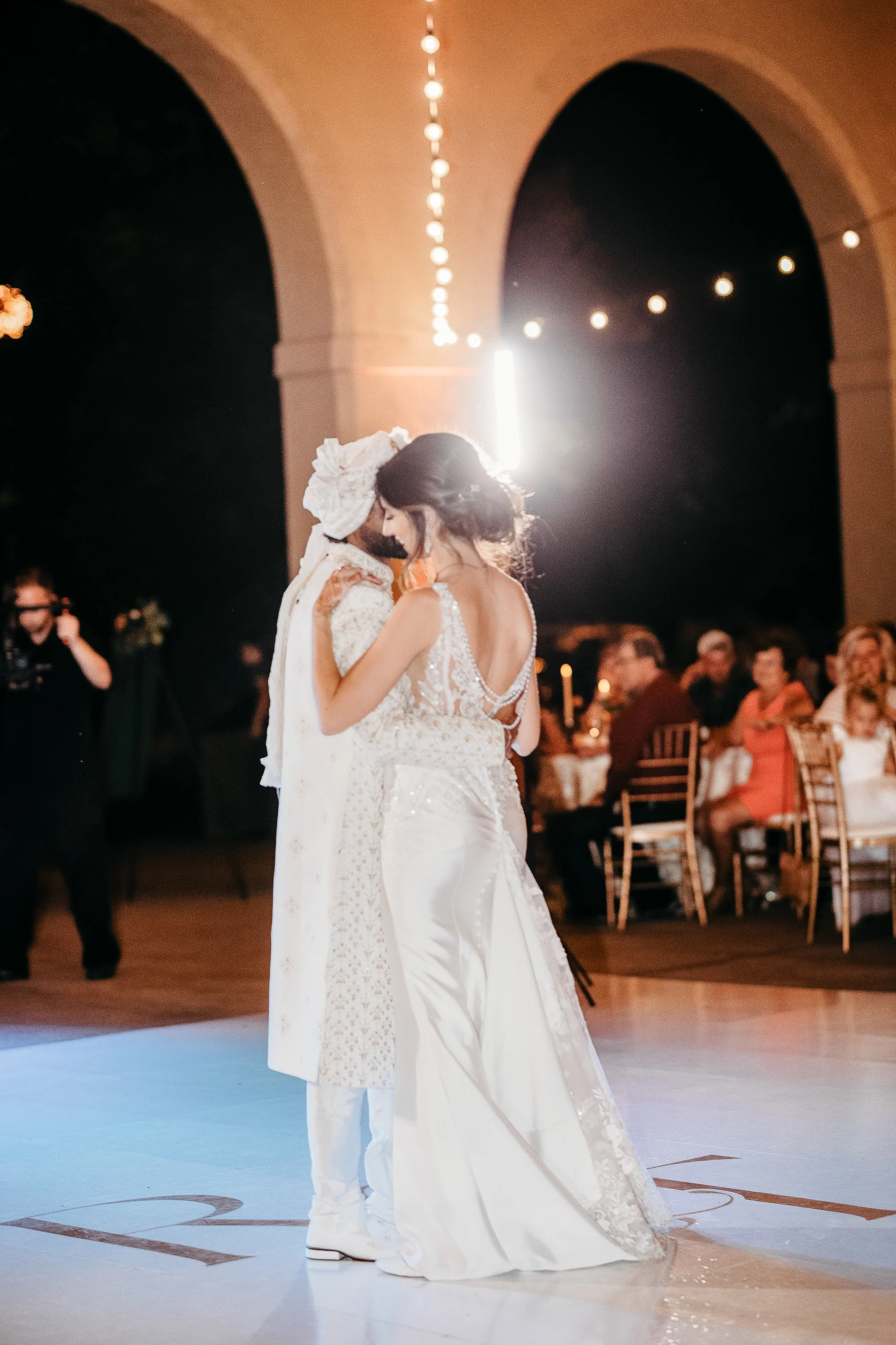 A bride and groom dancing at their wedding reception, surrounded by guests seated at tables, indoors with warm lighting and decorative arches.