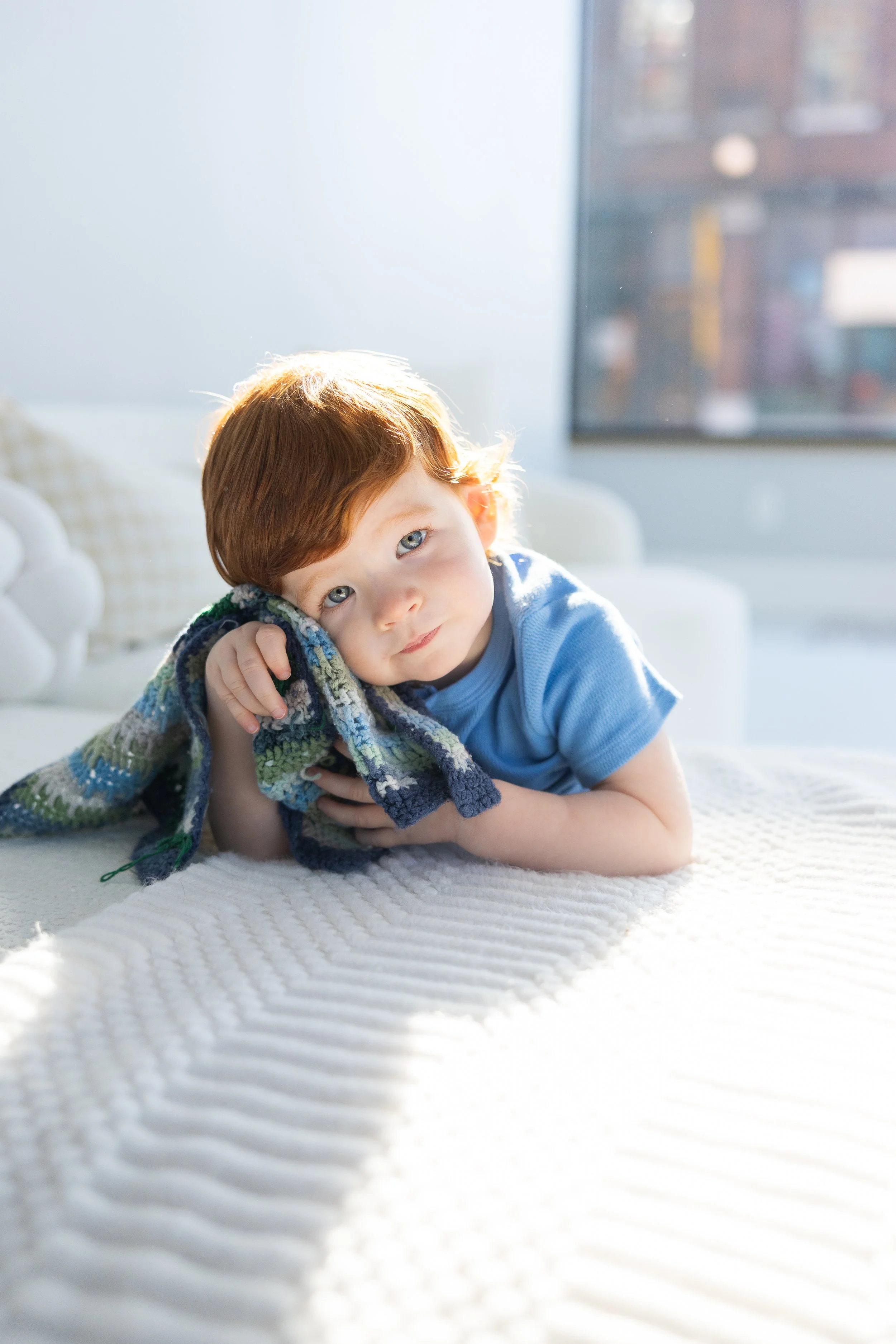 A young boy with red hair, lying on a white bed with a textured blanket, resting his head on a colorful knit blanket, with sunlight streaming through a nearby window.
