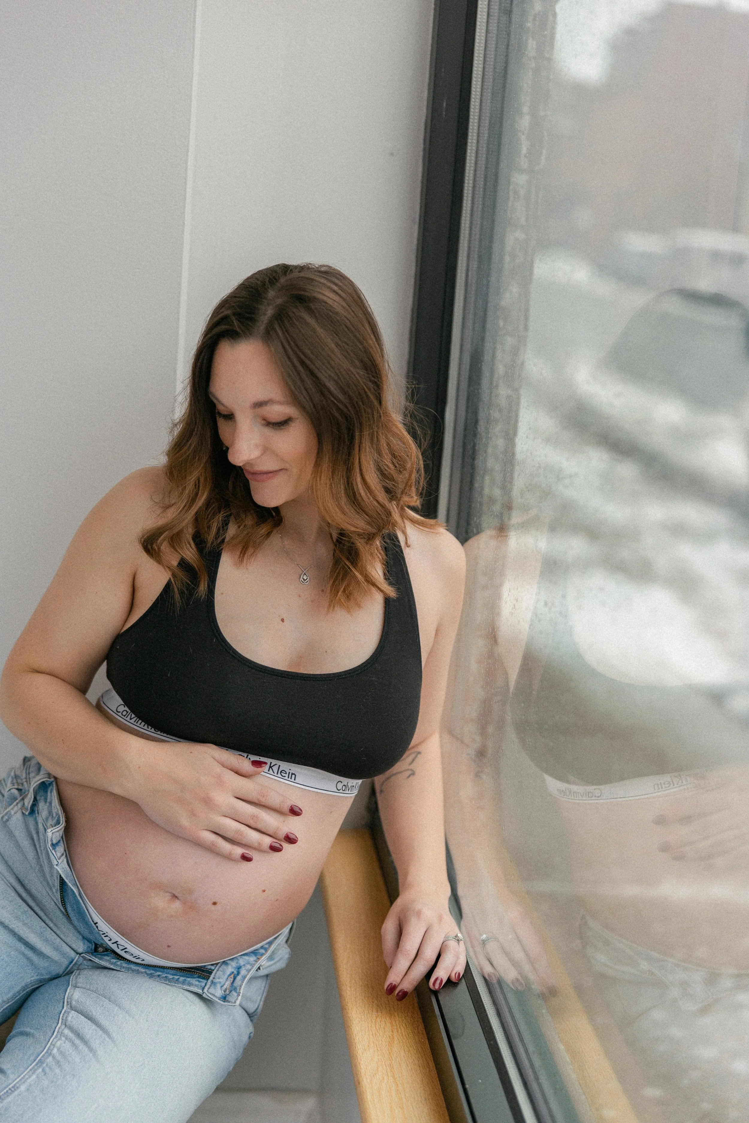 A pregnant woman with shoulder-length brown hair sitting by a window, looking down with a gentle smile, wearing a black Calvin Klein sports bra and light blue jeans.