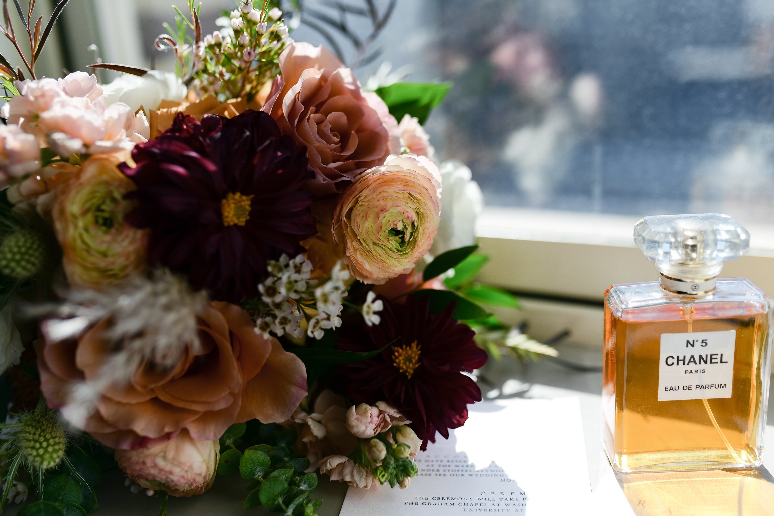 A bouquet of various flowers next to a bottle of Chanel No. 5 perfume on a windowsill, with a blurred background outside.