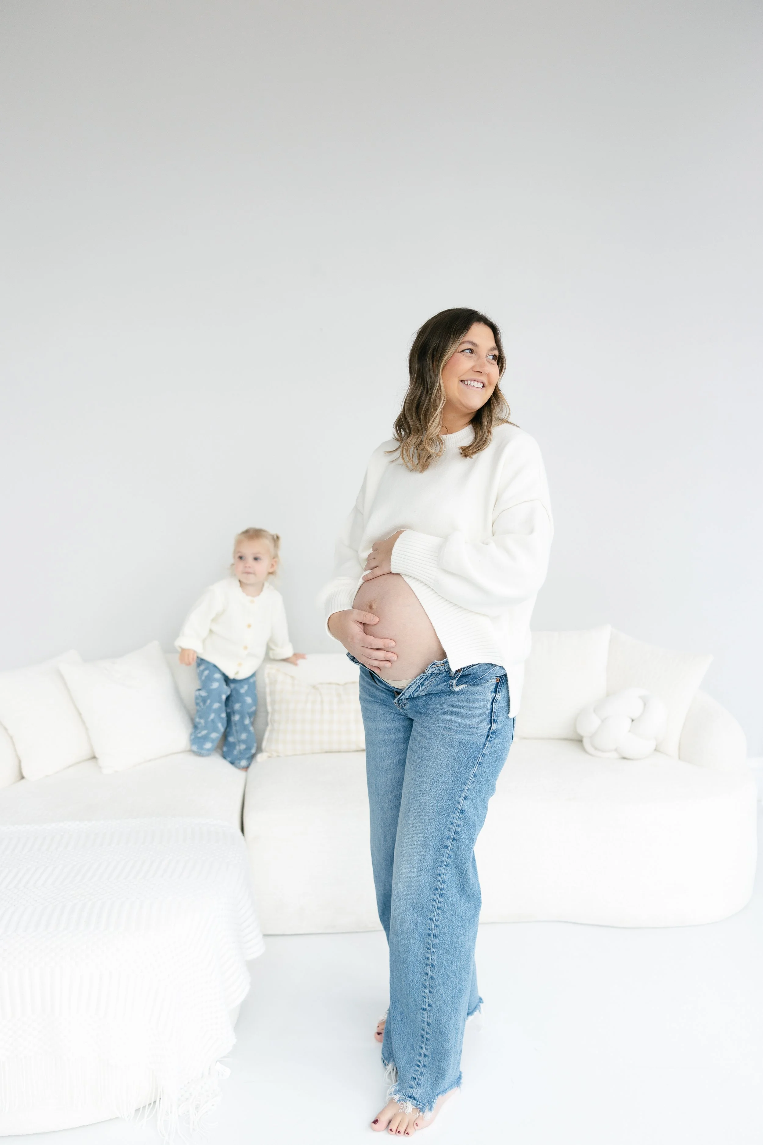 Pregnant woman standing in a white room, holding her belly with a smile, with a young girl sitting on a white sofa in the background.