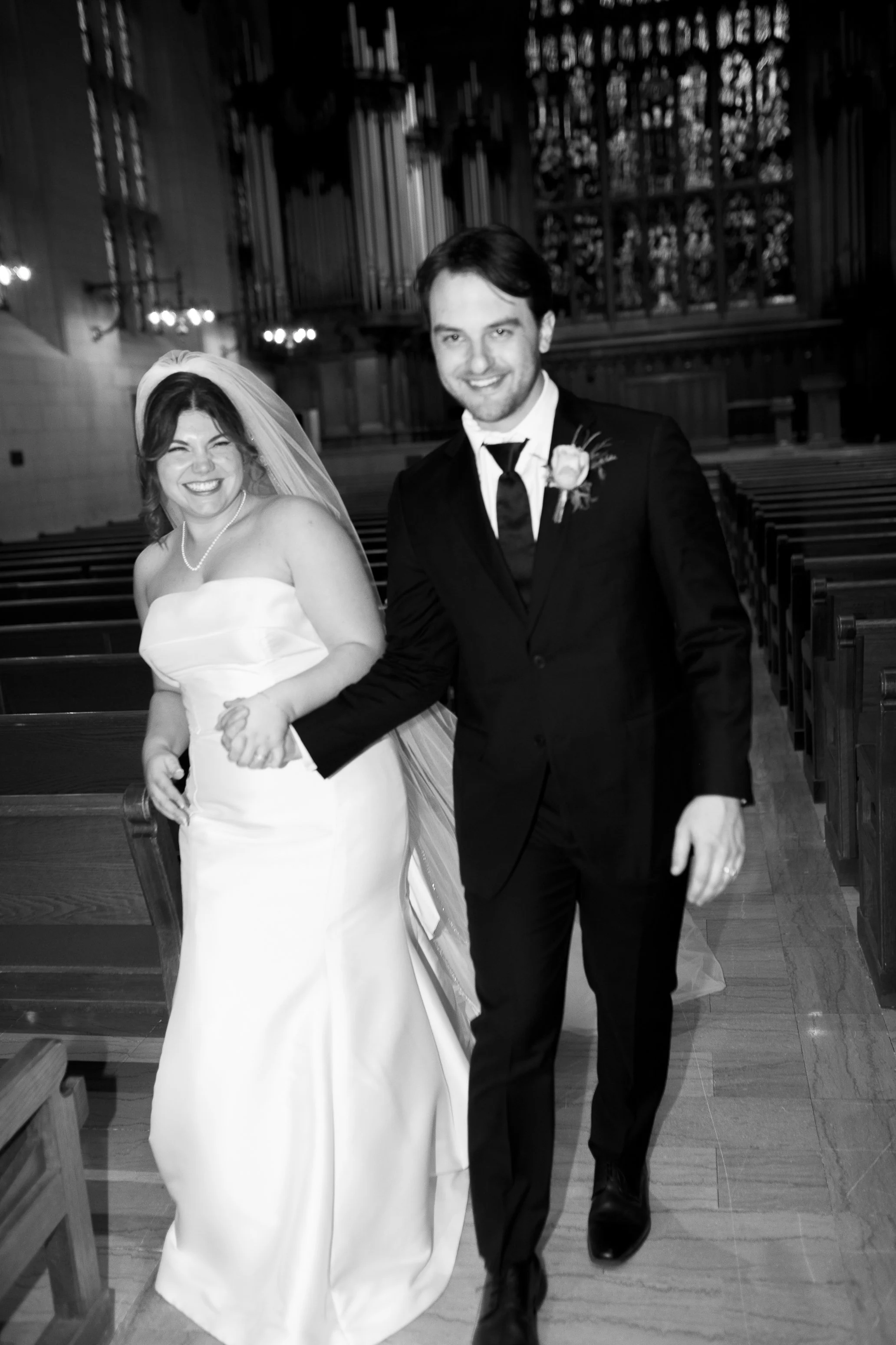 A bride and groom walking down the aisle in a church, smiling happily together. The bride is wearing a strapless wedding gown and a veil, the groom is in a dark suit with a boutonniere.
