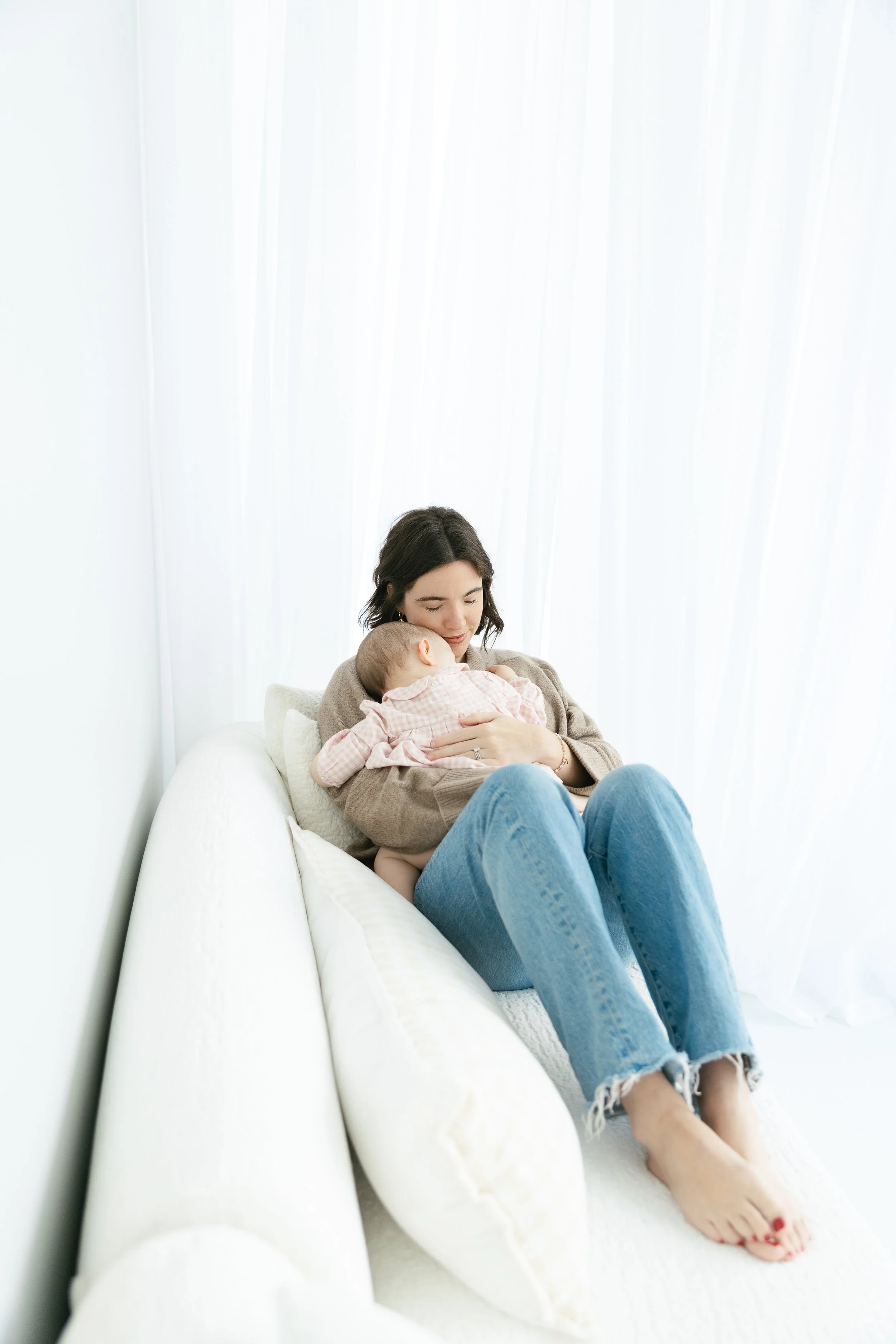 A woman with dark hair, wearing a brown sweater and blue jeans, sits on a white couch holding and breastfeeding a baby girl dressed in pink checked pajamas, with sunlight streaming through white curtains behind them.