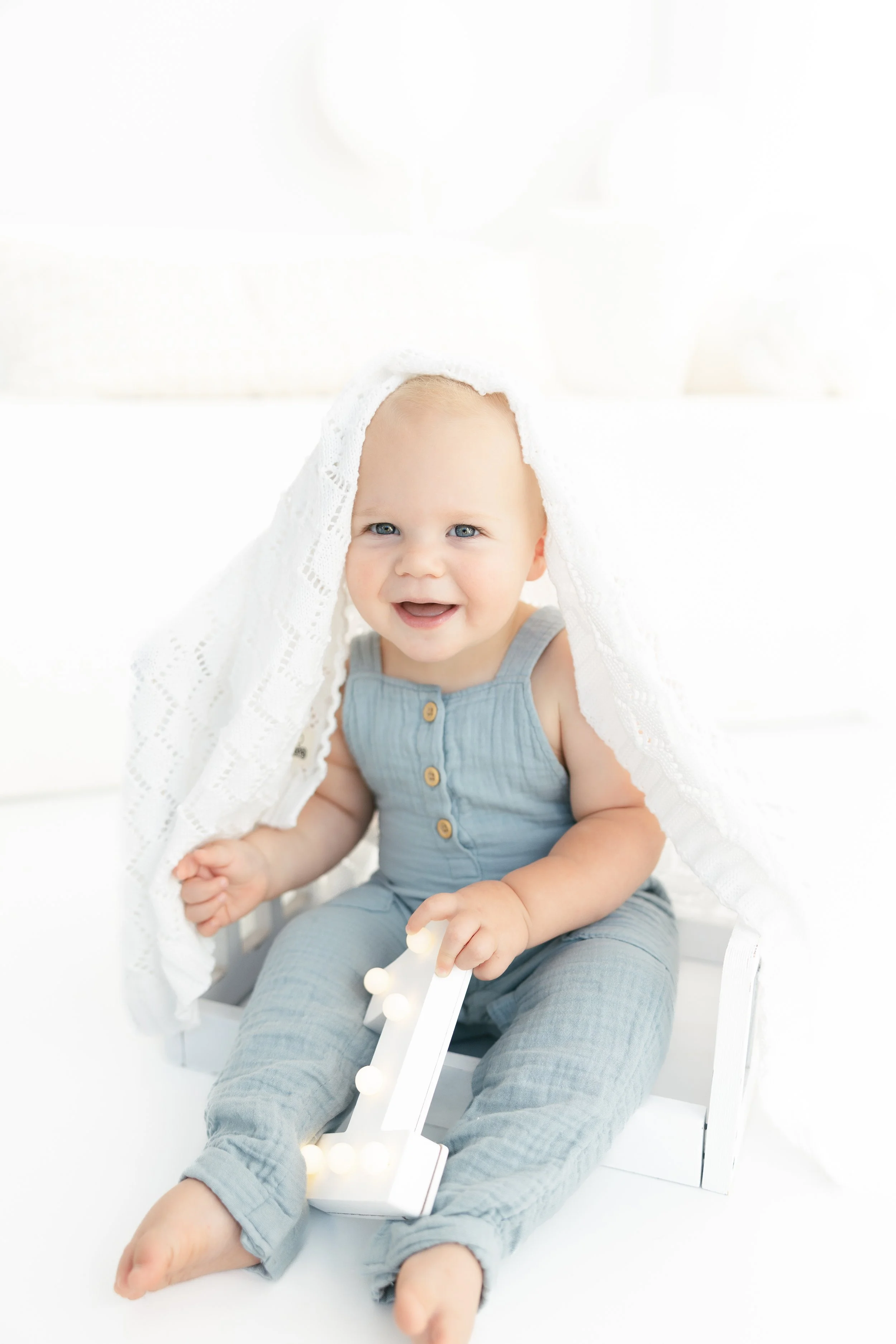 Smiling baby sitting on a white wooden chair, partially covered by a white lace cloth, holding a decorative lighted number one.