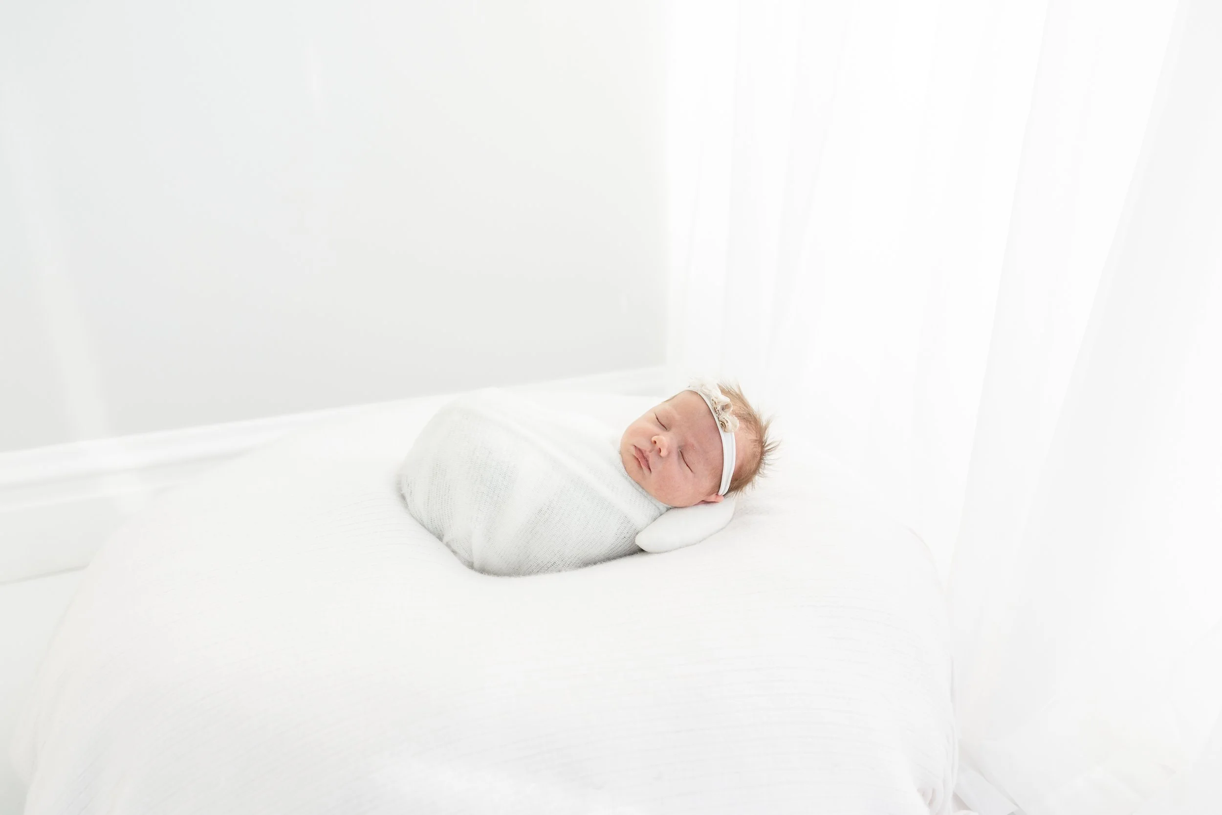 A newborn baby bundled in white swaddle, sleeping on a white bed with a white wall in a bright room.