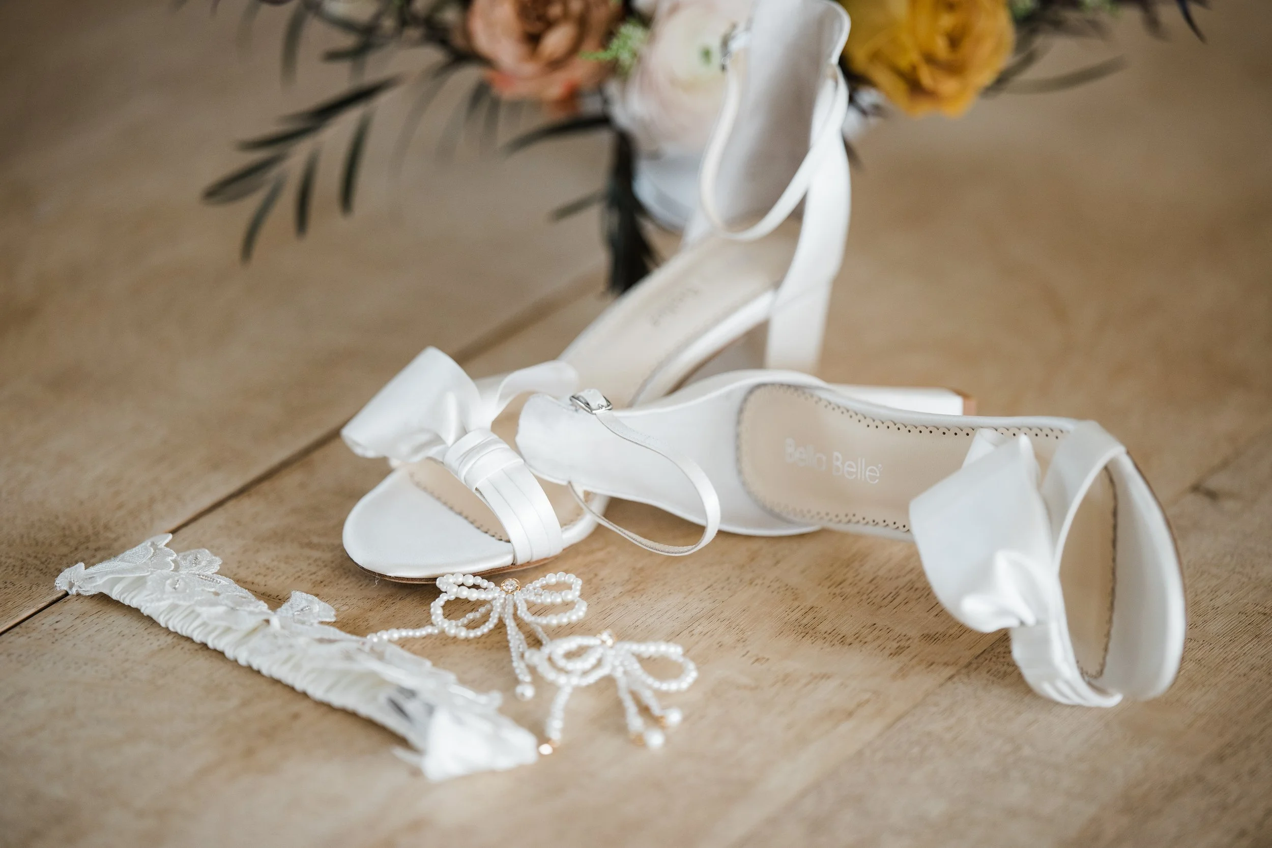 A pair of white high-heeled shoes with bows, a pearl necklace with bow charms, and a fabric headband with lace and ribbon details are displayed on a wooden surface, with a floral arrangement in the background.