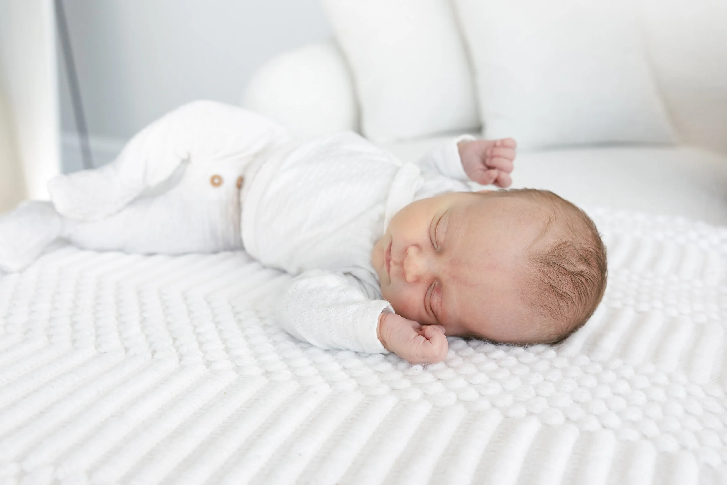 A sleeping newborn baby lying on a white textured blanket on a bed, dressed in a white outfit.