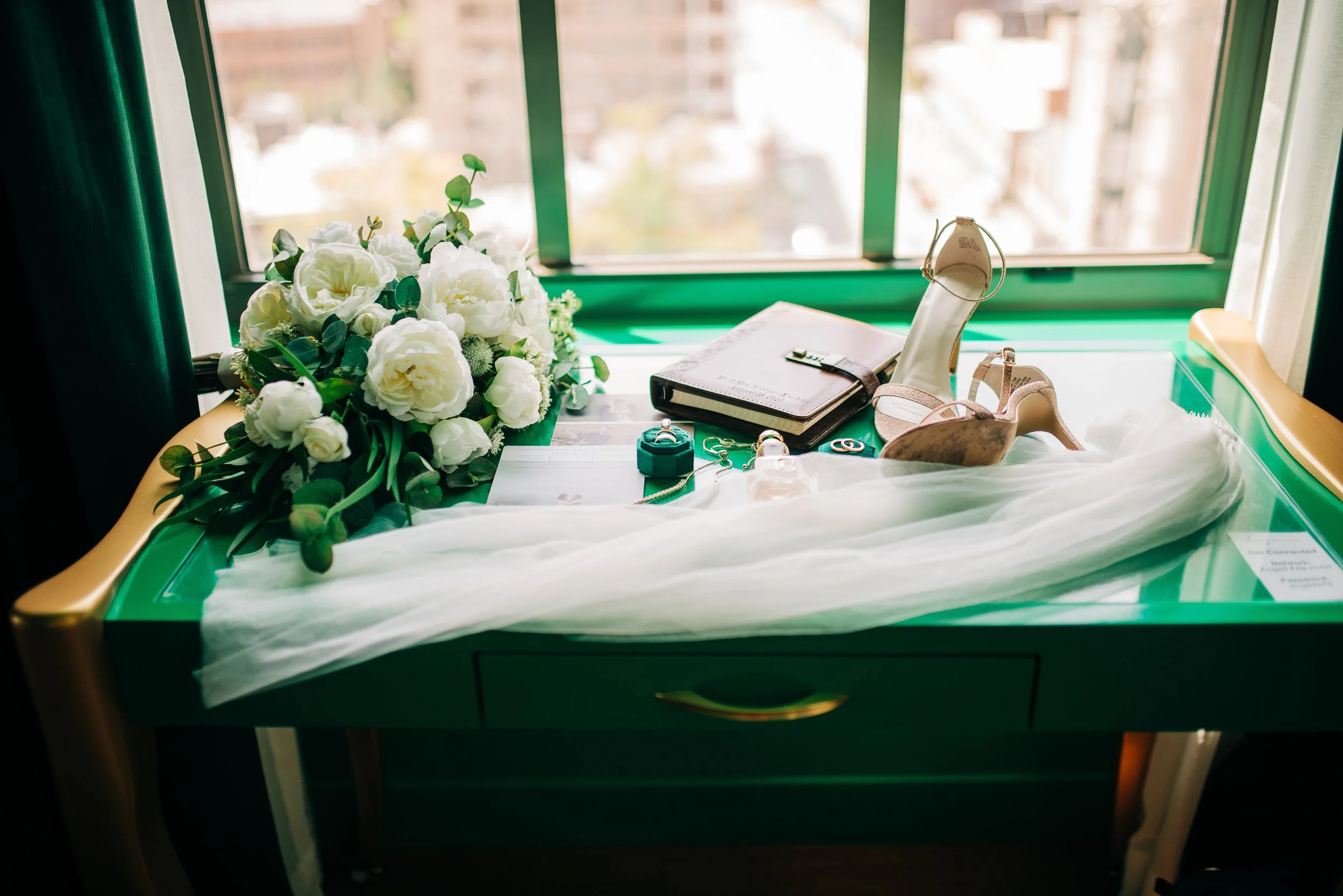 Wedding accessories on a green table near a window, including a bouquet of white flowers, a wedding dress veil, high-heeled shoes, a notebook, jewelry, and other small accessories.