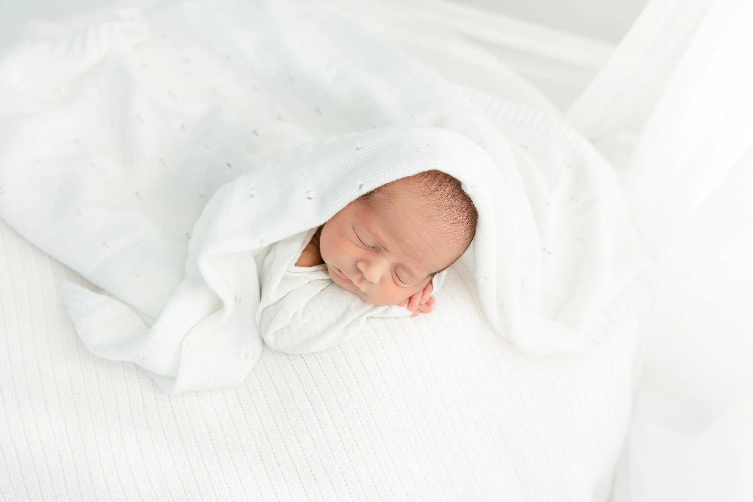 Newborn baby sleeping peacefully, wearing a white knit hat and wrapped in a white blanket, lying on a white textured surface.