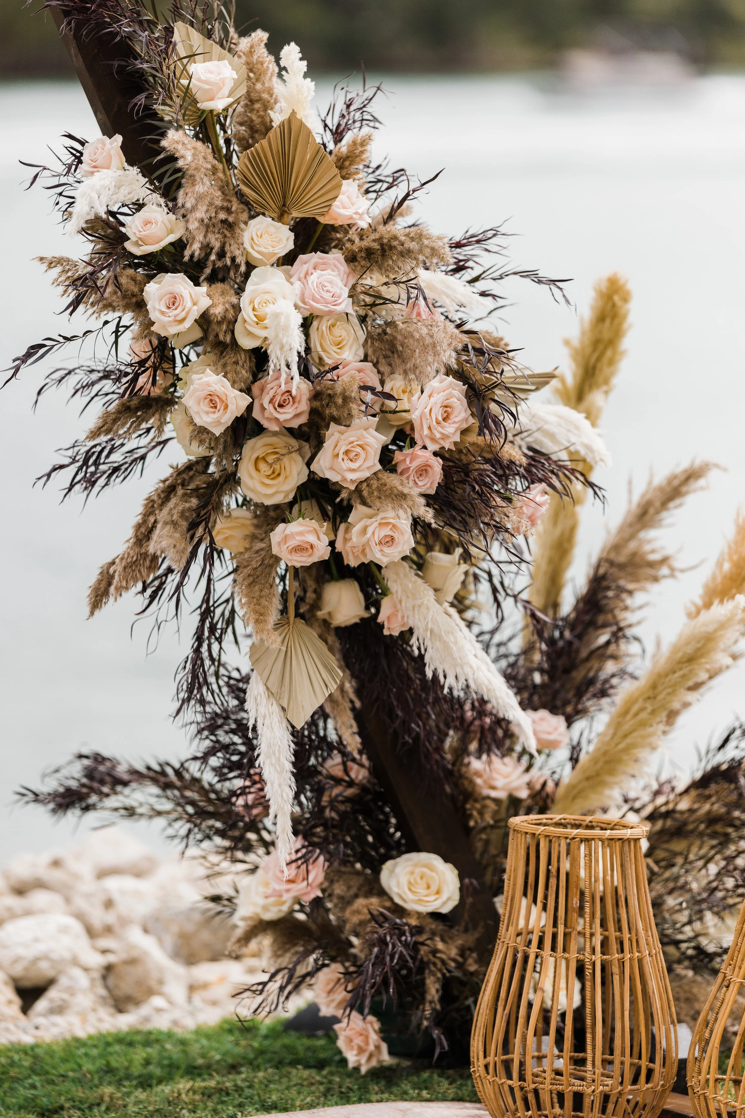 A floral arrangement featuring pale pink and white roses, dried pampas grass, and brown leaves, near a body of water with rocks and grass in the background.