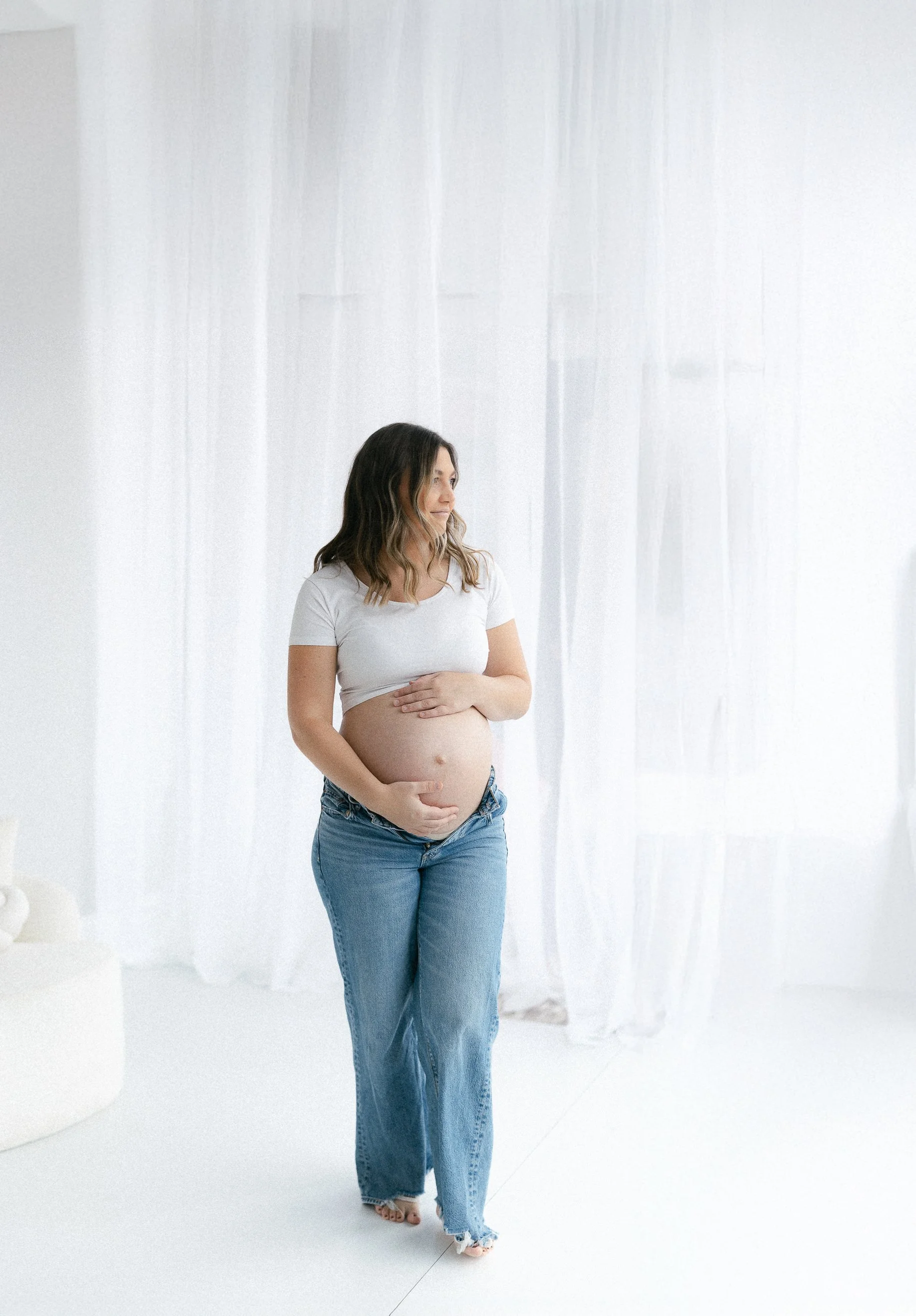 A pregnant woman standing in a bright, white room with sheer curtains, wearing a white t-shirt and blue jeans, touching her belly and looking to the side.
