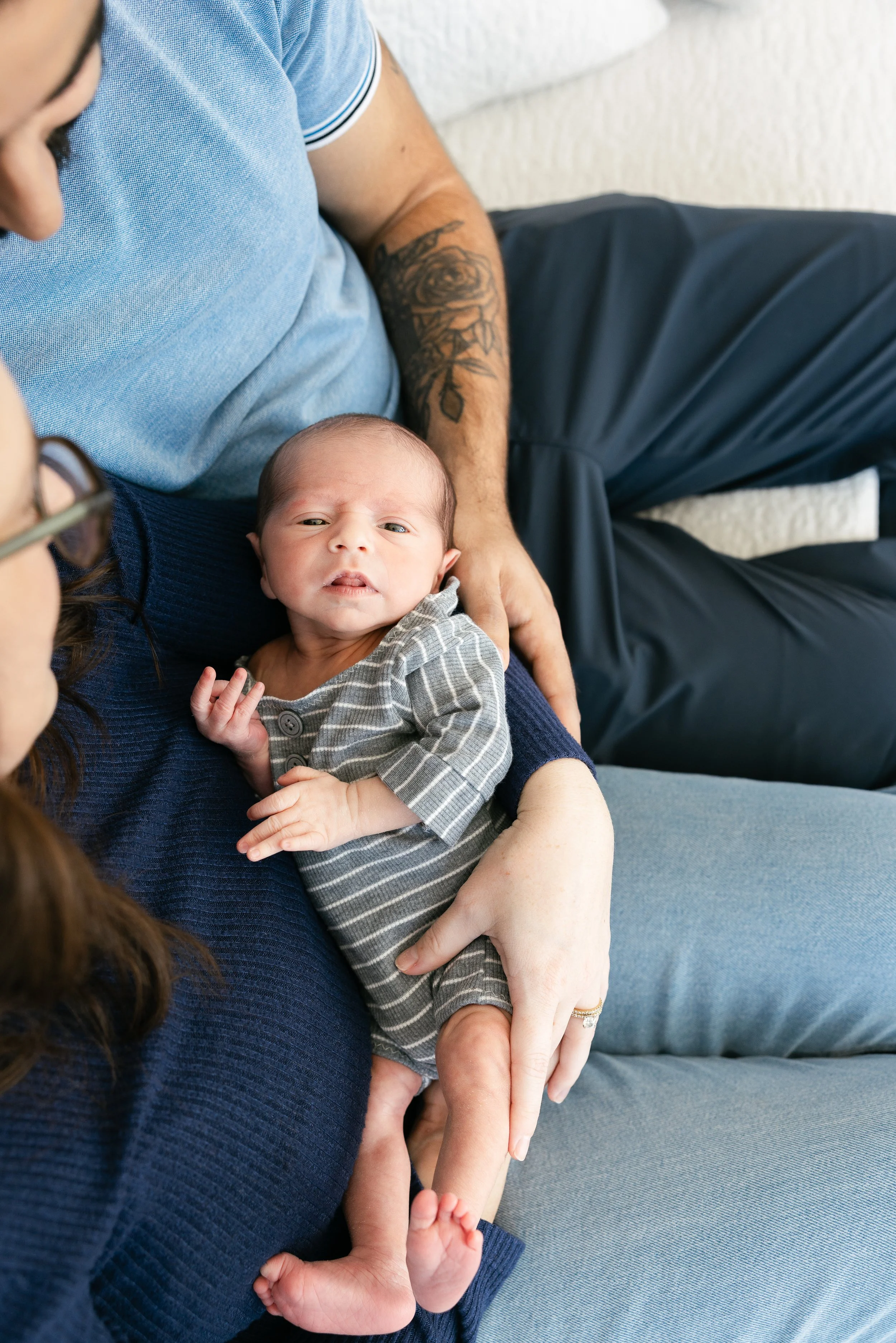 A newborn baby resting on a woman's lap, with a man sitting next to them. The baby is wearing a striped gray onesie and looks at the camera with a curious expression.