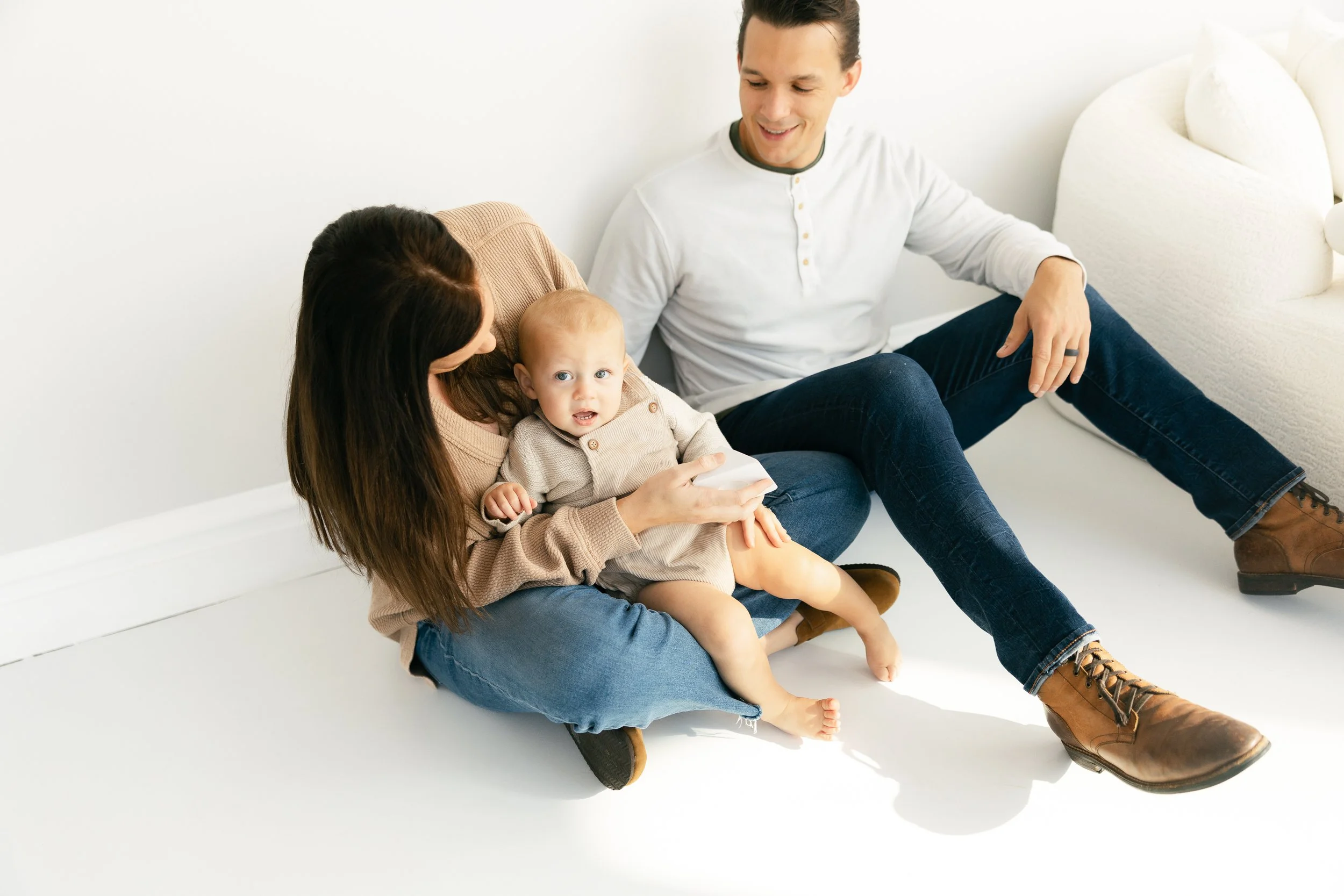 A family sitting on the floor, consisting of a woman holding a young boy, a man smiling, and a white sofa in the background.