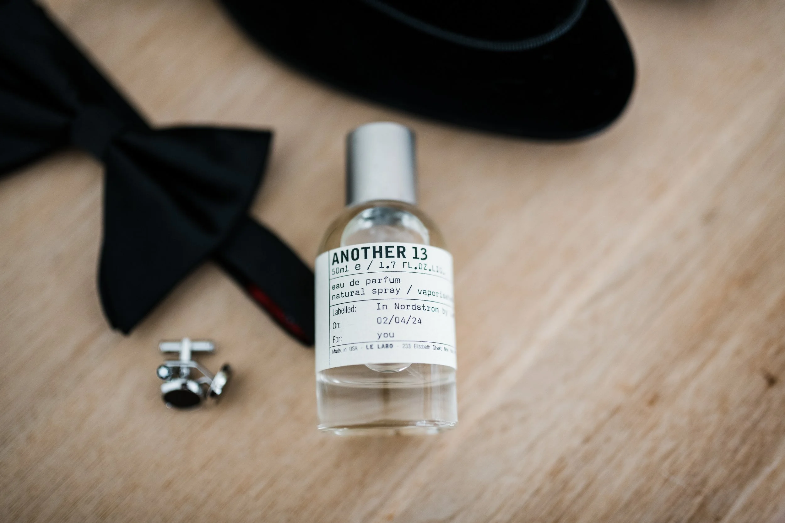 A small bottle of perfume labeled 'Another 13', a black bow tie, a metallic cufflink with a black face, and a black object in the background on a wooden surface.