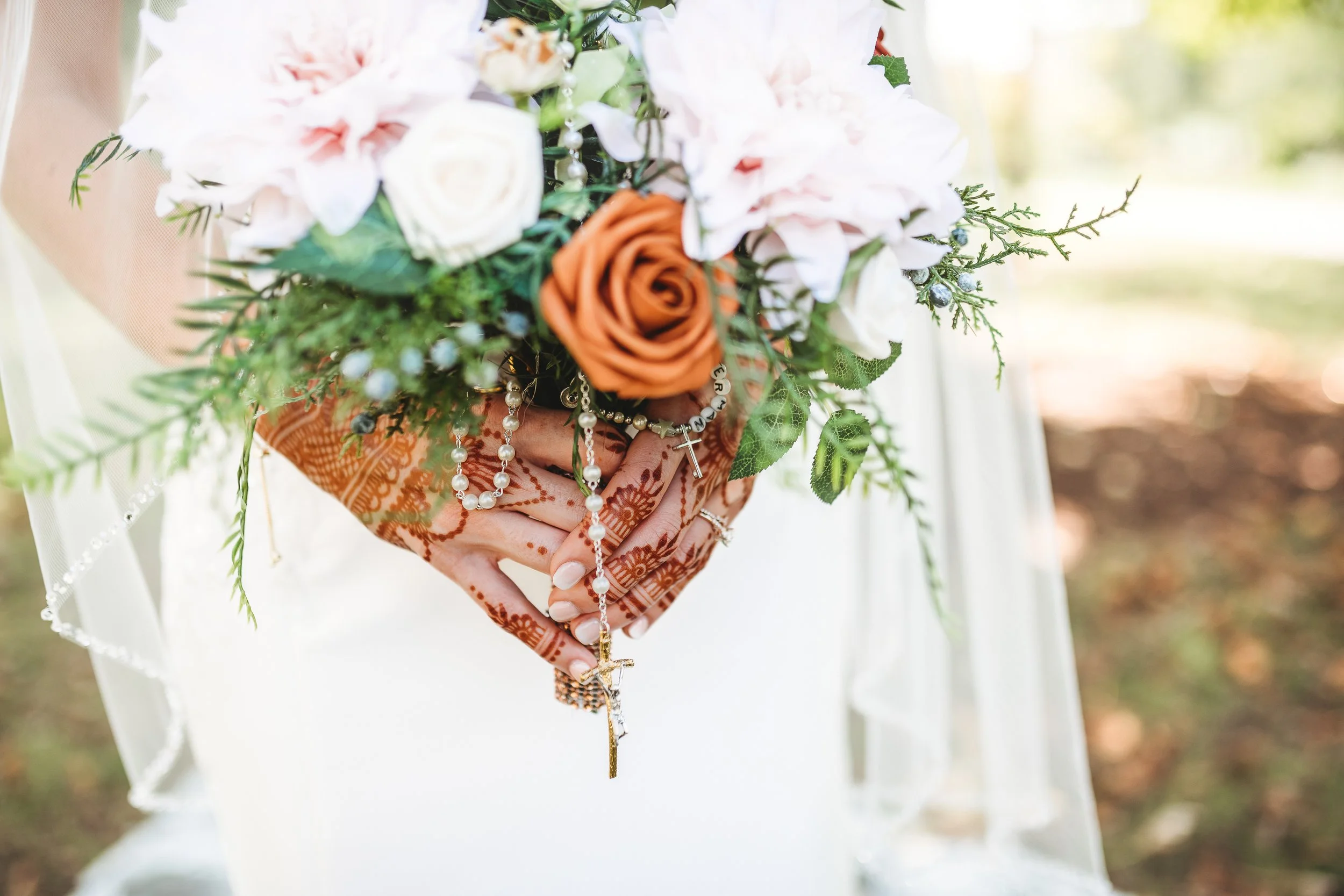 A bride holds a bouquet of white and orange roses, roses, and greenery, with a rosary hanging from her hands. The bride's hands have henna designs.