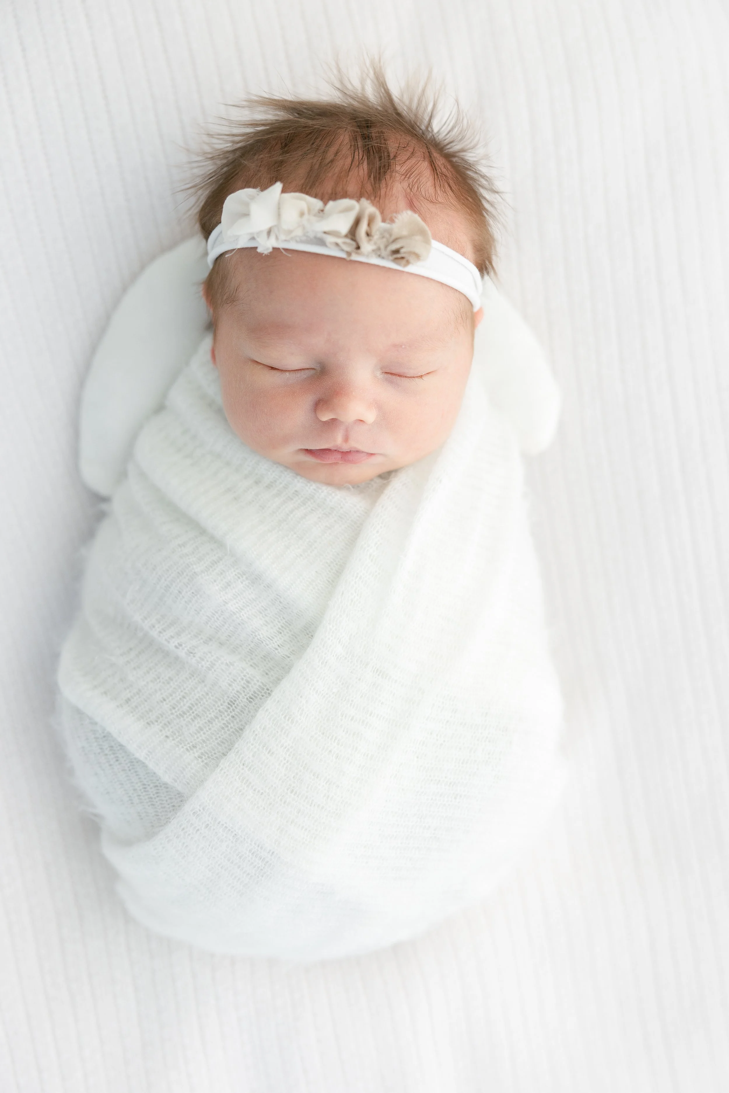 Close-up of a sleeping newborn baby wrapped in a white blanket, wearing a cream-colored headband with fabric flowers.