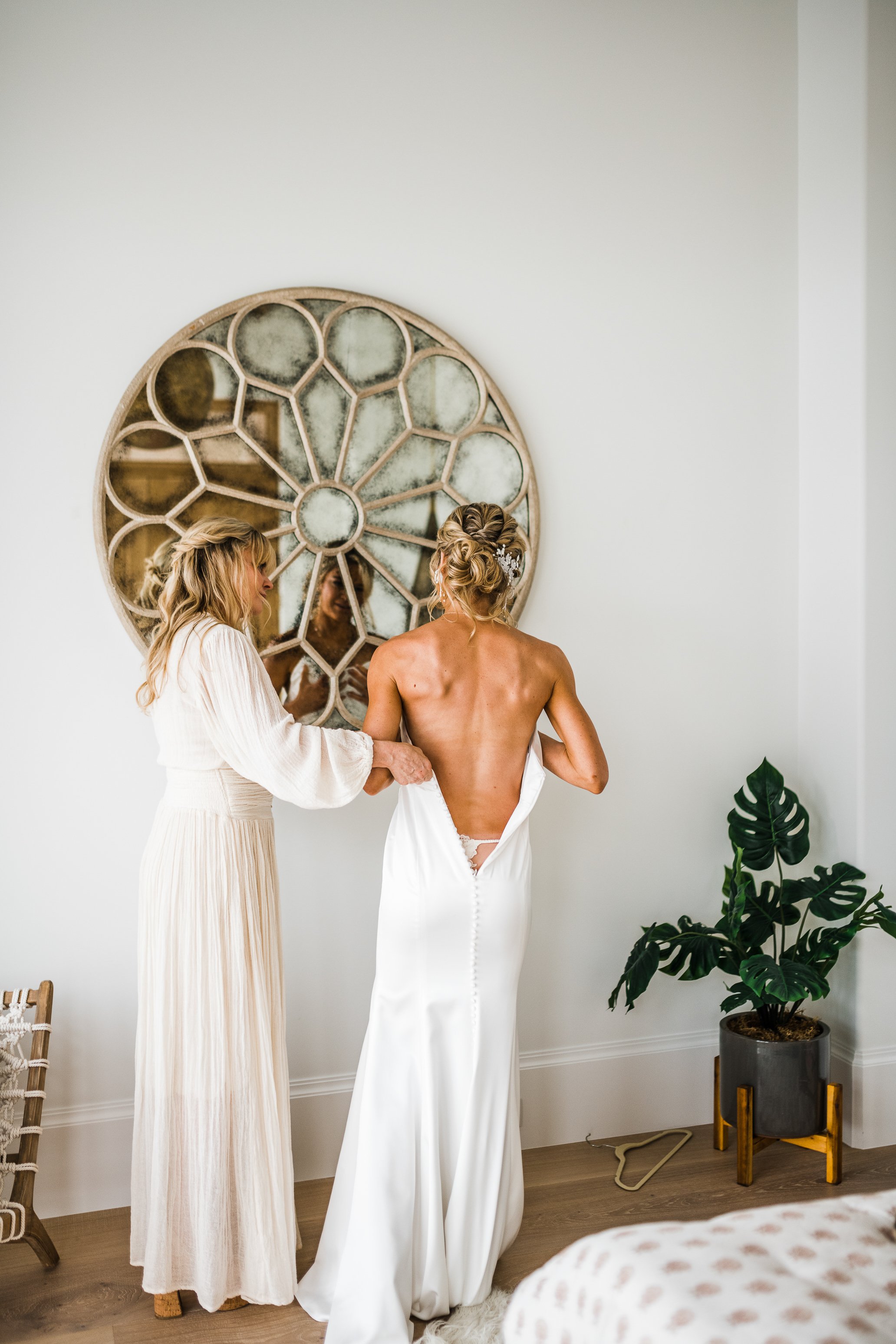 A bride in a white wedding dress with a deep back is being assisted by another woman, possibly her mother or a stylist, in putting on or adjusting her dress inside a bright room with a large decorative mirror and a potted plant.