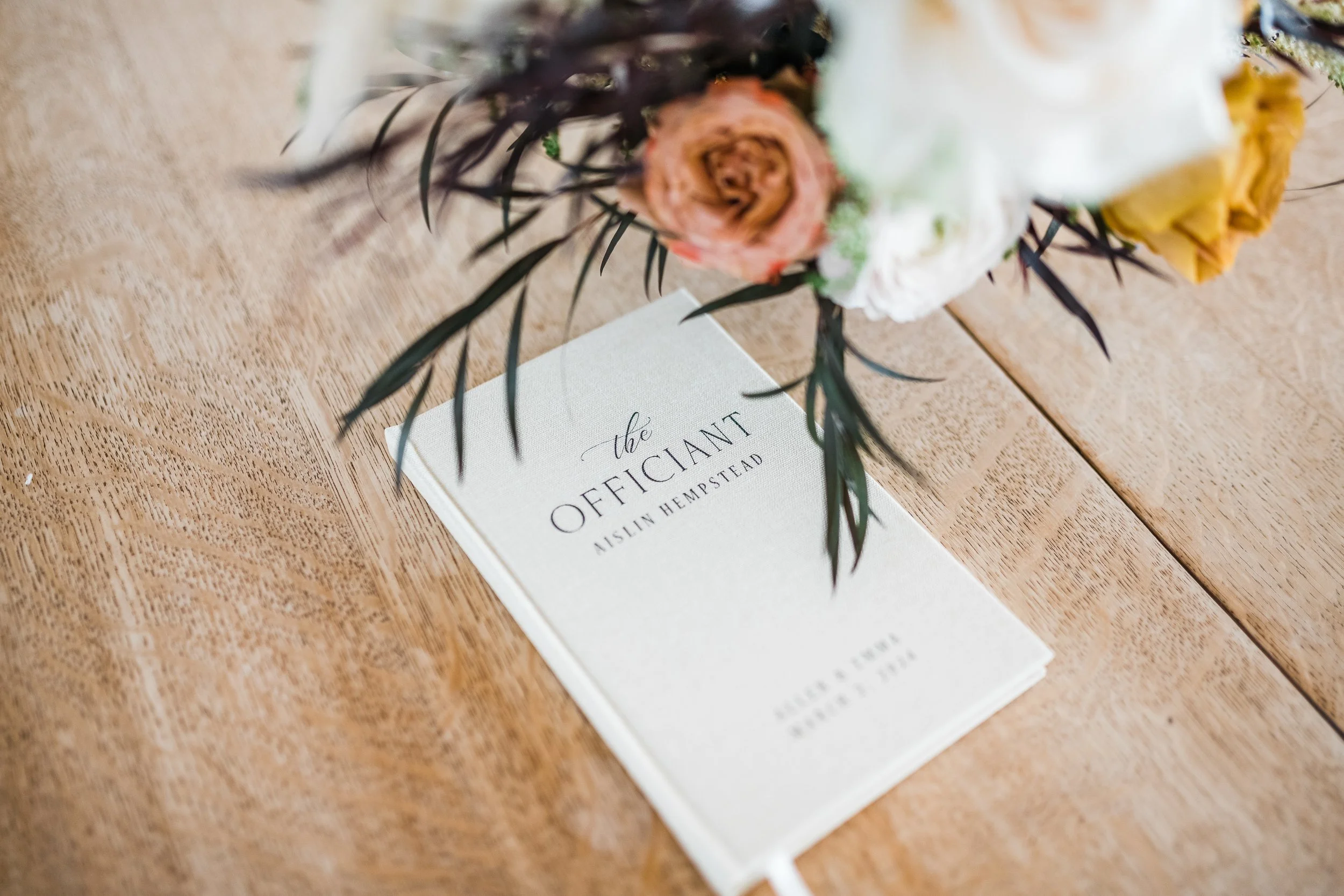 A bouquet of flowers resting on a wooden table, partially covering a white booklet titled 'The Officiant' by Nislin Hempstead.