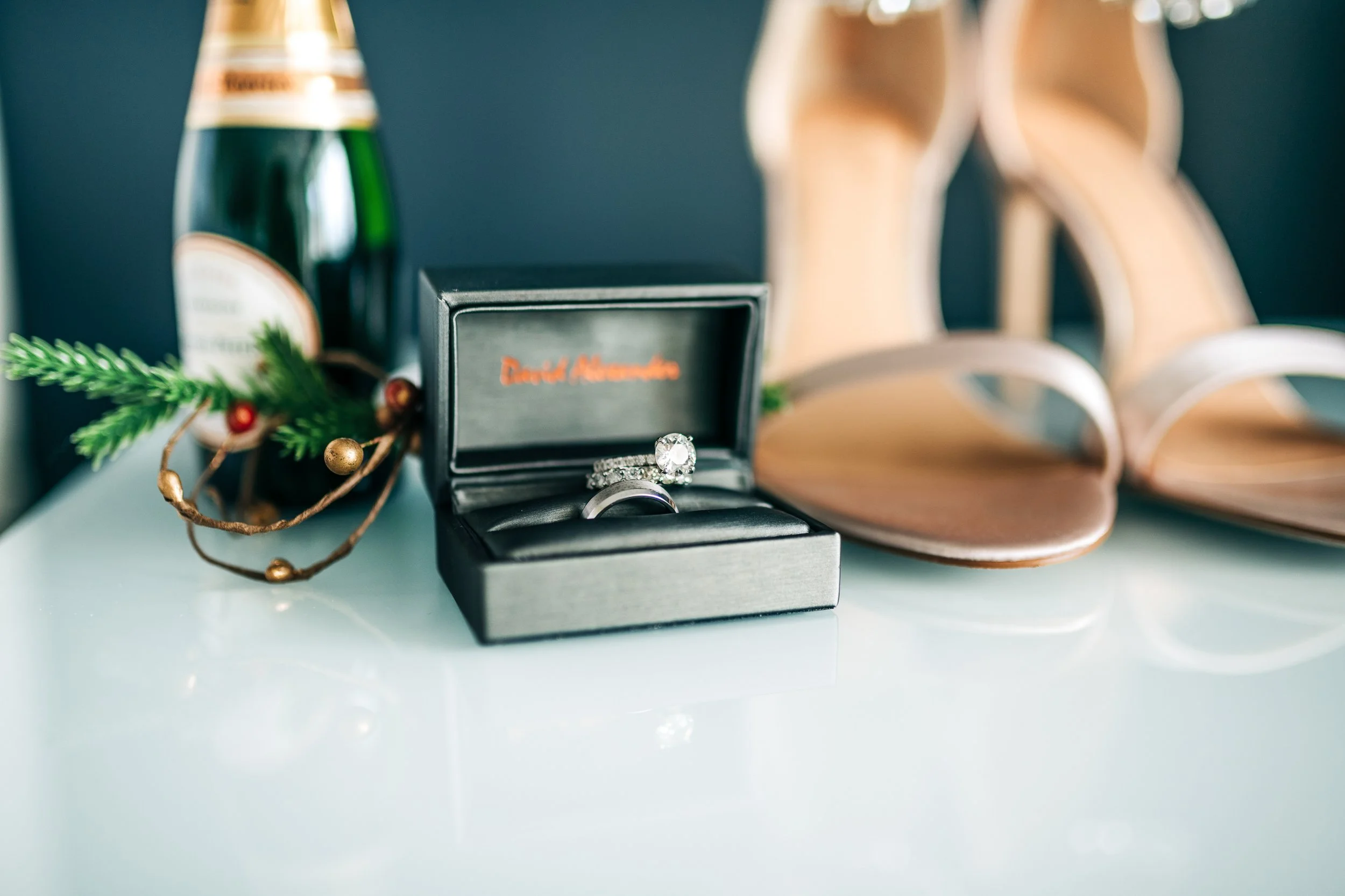 A black jewelry box with a diamond engagement ring inside, surrounded by brown high heel shoes, a Chanel bottle, and holiday decorations.