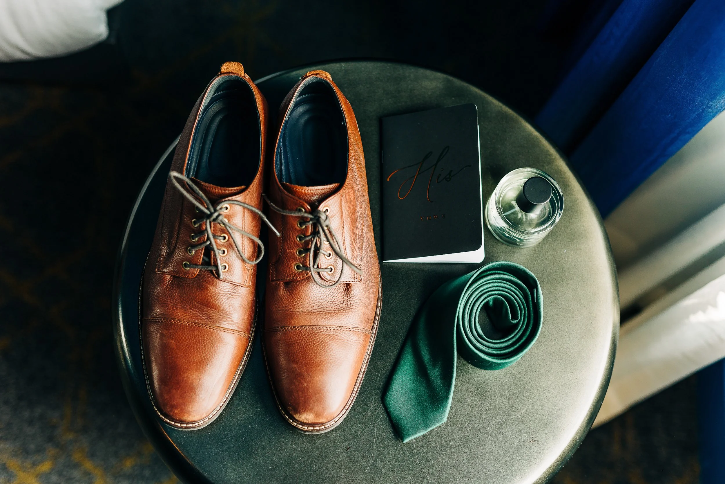 Brown leather dress shoes, green tie, black notebook labeled "His", perfume bottle, and rolled-up green sock on a small round table near a window.