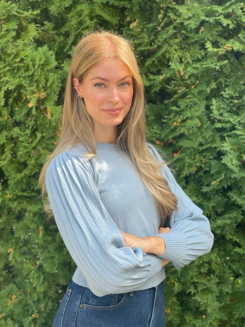 A young woman with blonde hair, blue eyes, wearing a light blue sweater with pleated sleeves and dark jeans, standing outdoors in front of lush green foliage with crossed arms, looking at the camera.