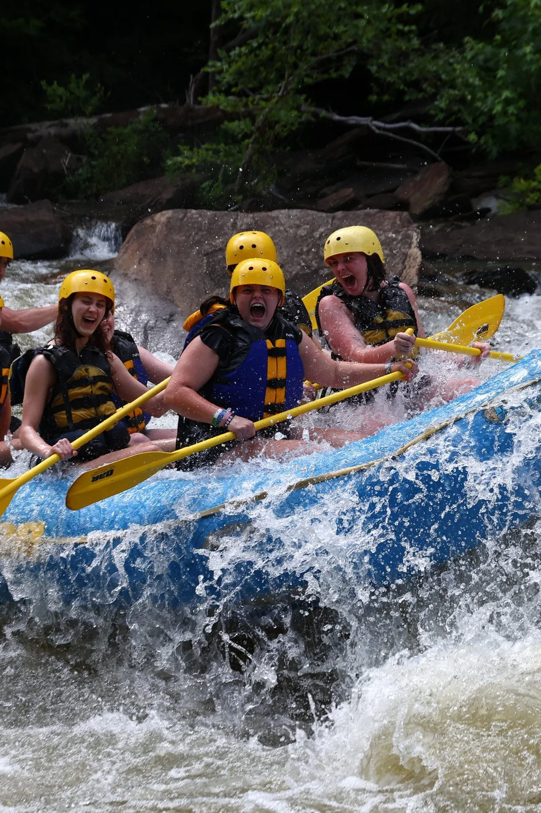 Group of people white water rafting on a river, wearing yellow helmets and life jackets, holding paddles with excited expressions.