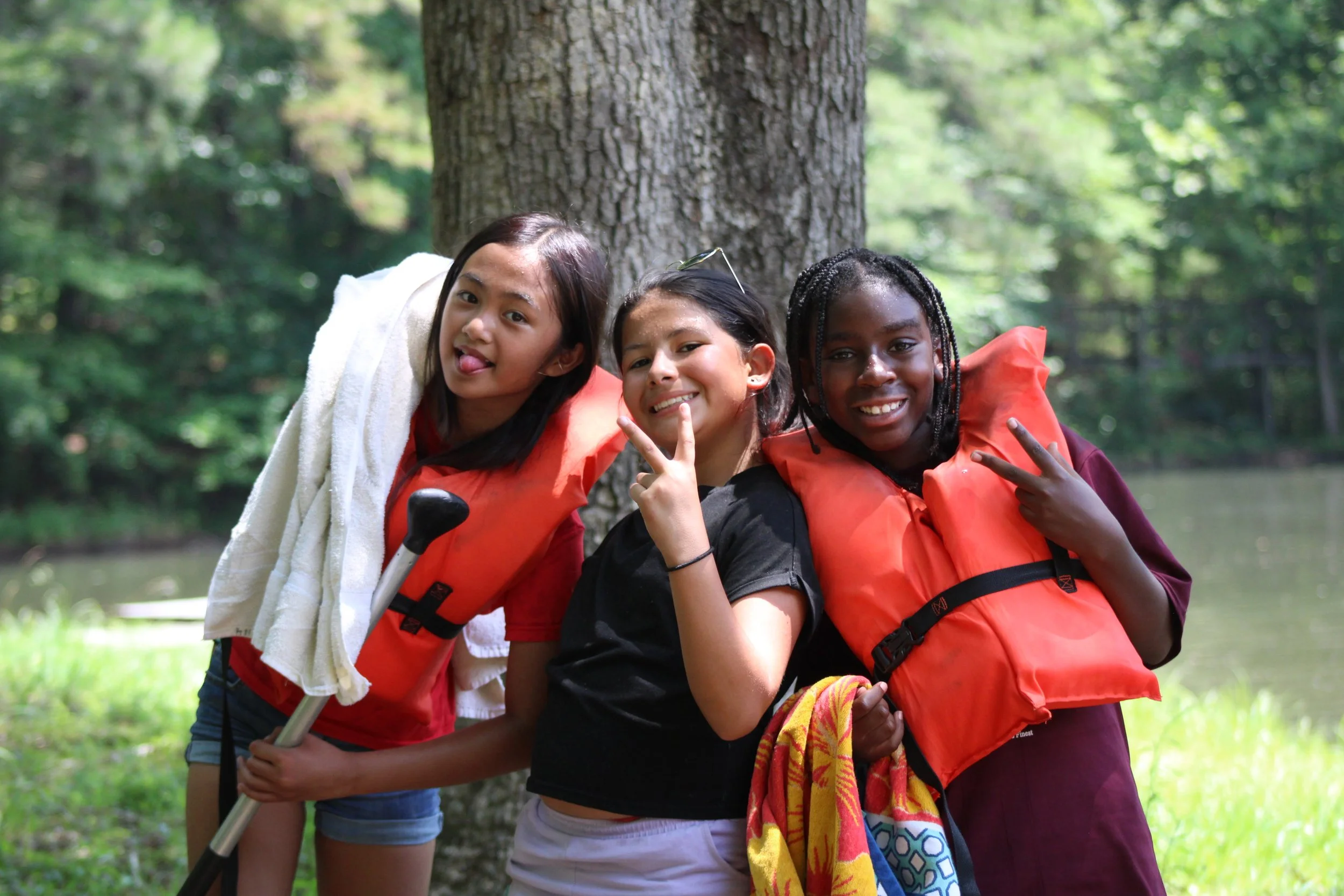Three girls smiling and posing in front of a tree in a forest, wearing life jackets.