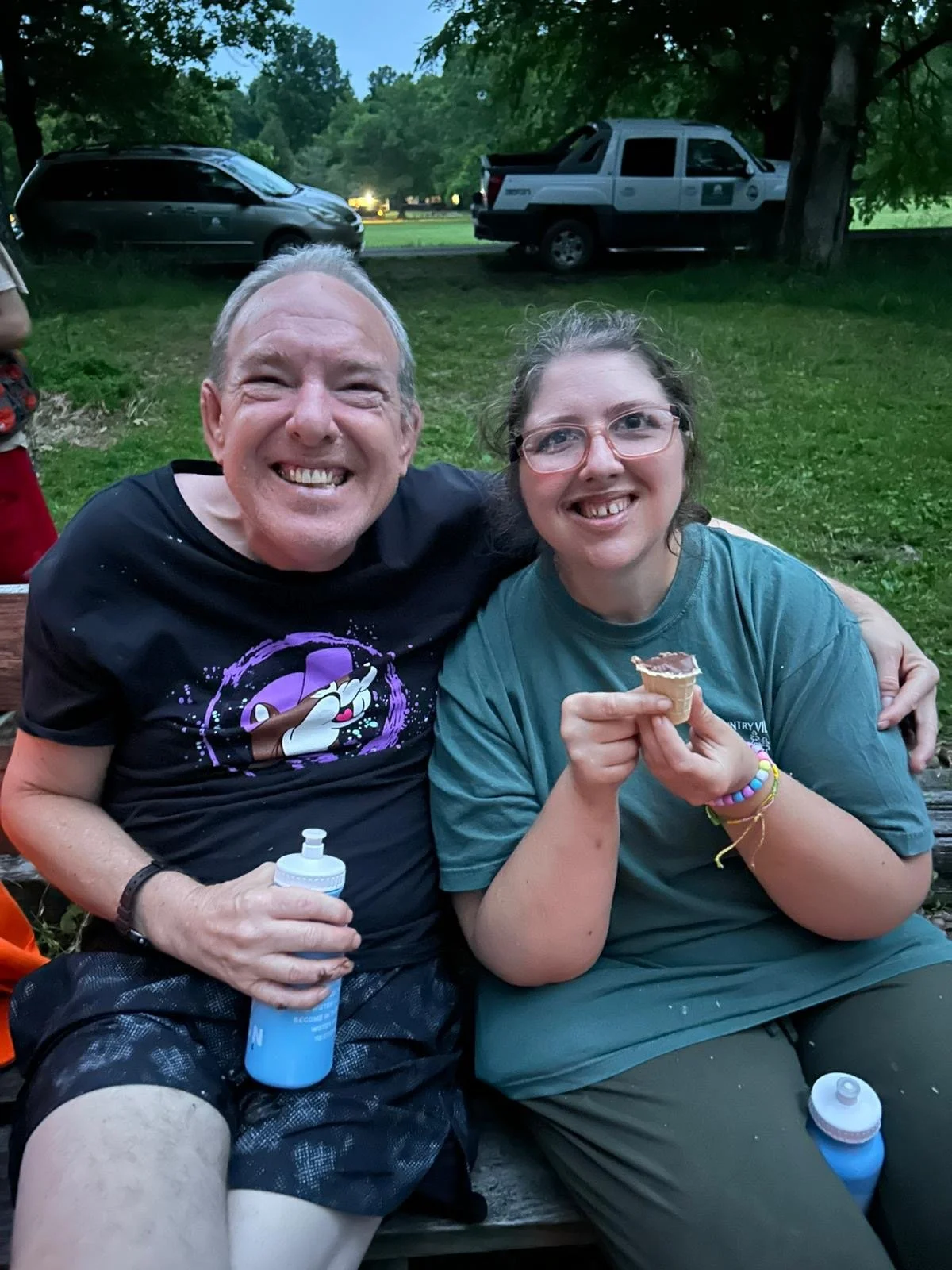 Two smiling people sitting on a park bench outdoors, with cars in the background. The person on the left is an older man holding a water bottle, and the person on the right is a young woman holding a small ice cream cone with some chocolate on top.