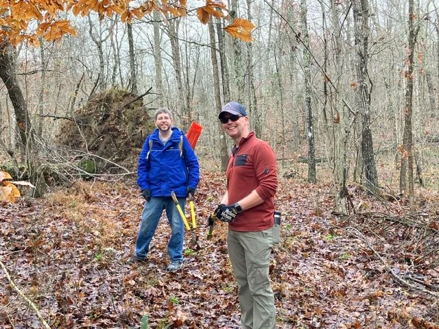 Two men hiking in a forest with leafless trees and fallen leaves on the ground.