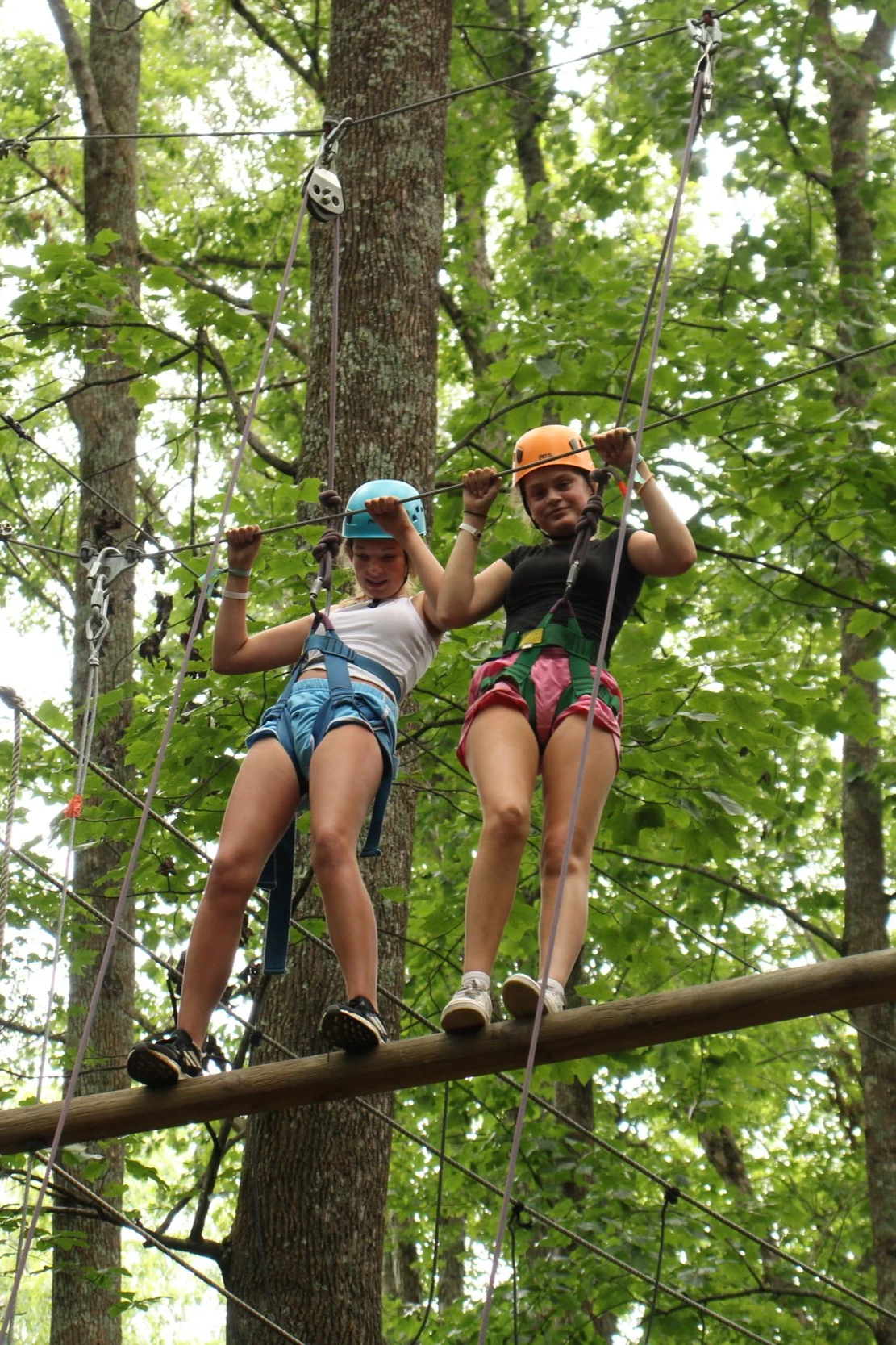 Two girls wearing helmets and harnesses walking on a wooden crosswalk between trees in a forest