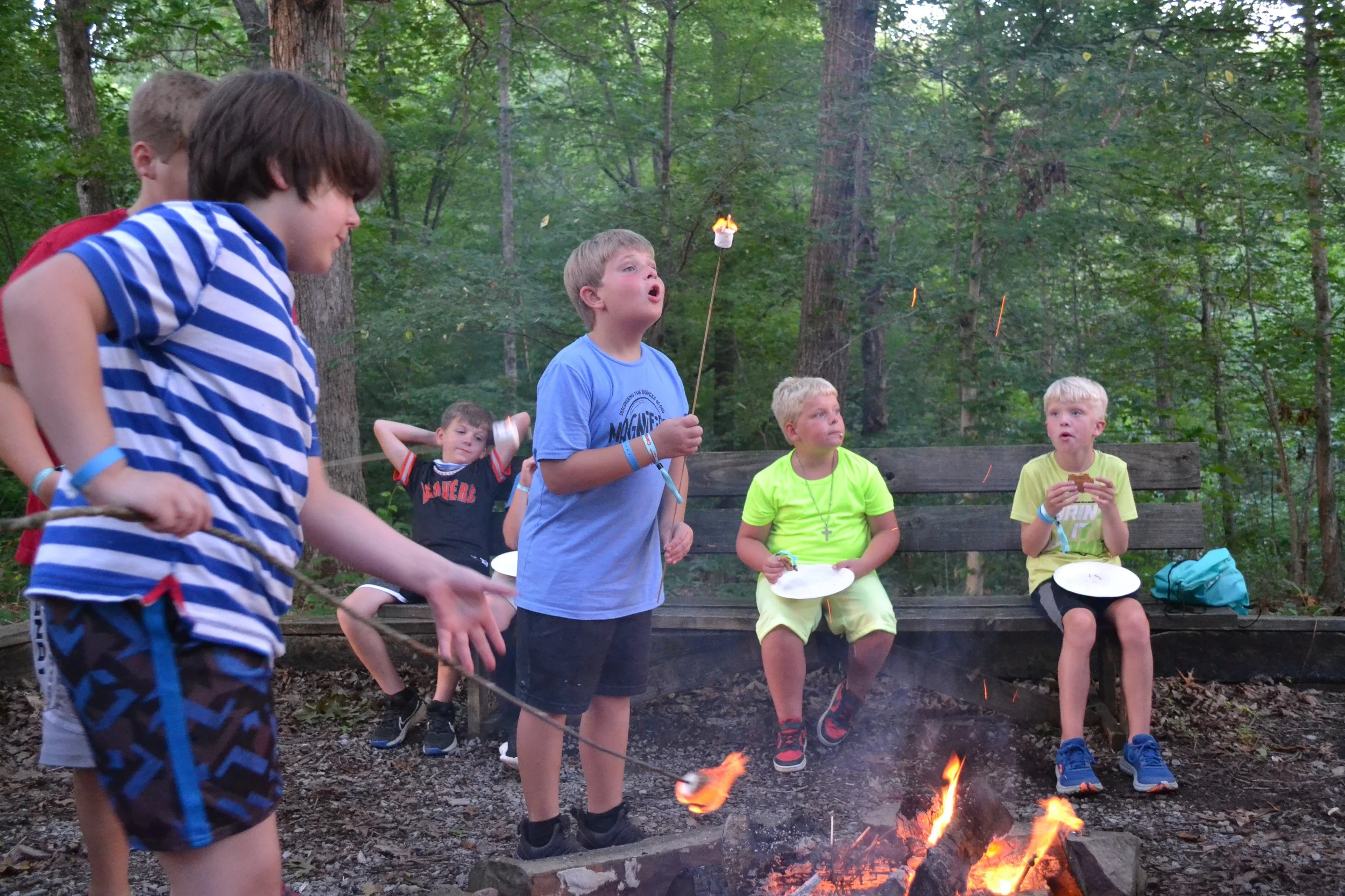 Children sitting around a campfire in a wooded area, some roasting marshmallows and eating snacks during a campfire activity.