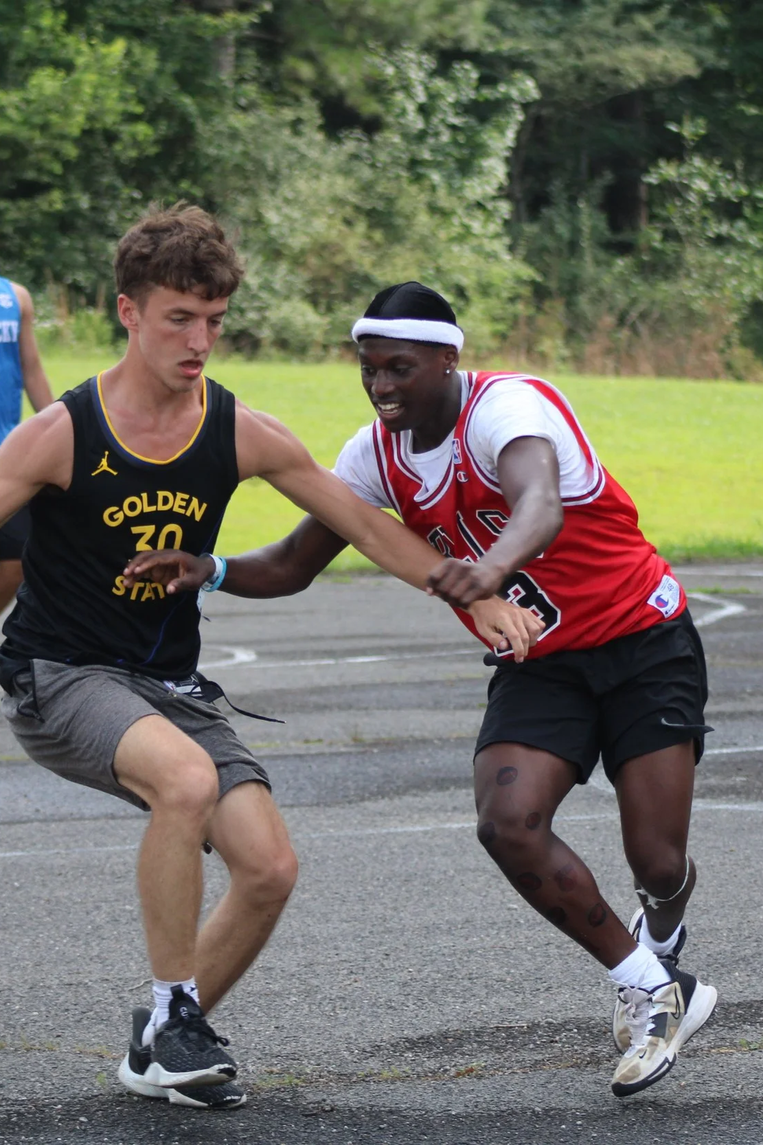 Two young men are running and competing in a race, with one wearing a black tank top and the other wearing a red sports jersey, on an outdoor track, with greenery in the background.