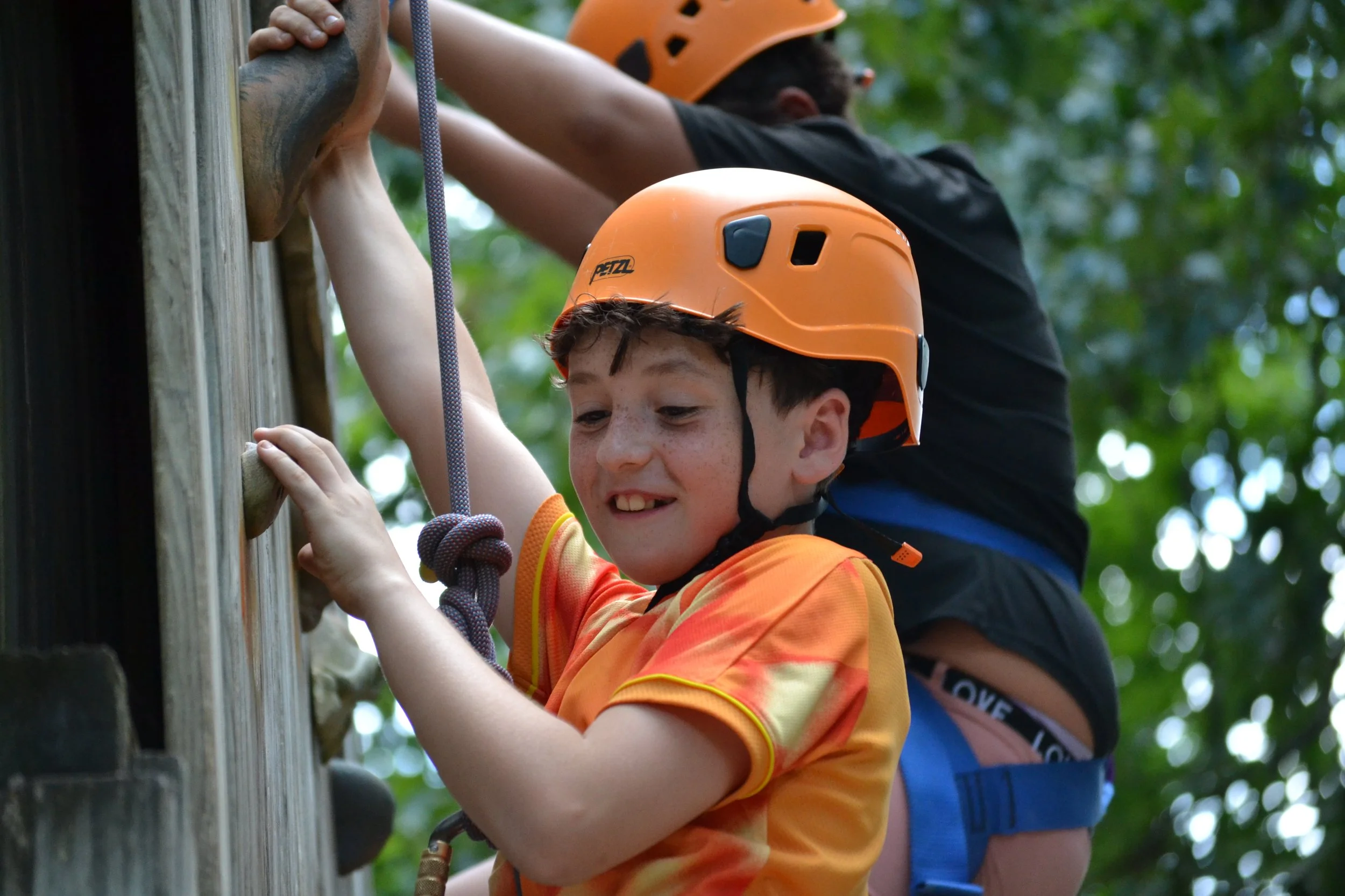 A young boy wearing an orange helmet and orange sports jersey, smiling as he climbs a wooden rock wall with the help of an adult behind him. The background shows green leafy trees.