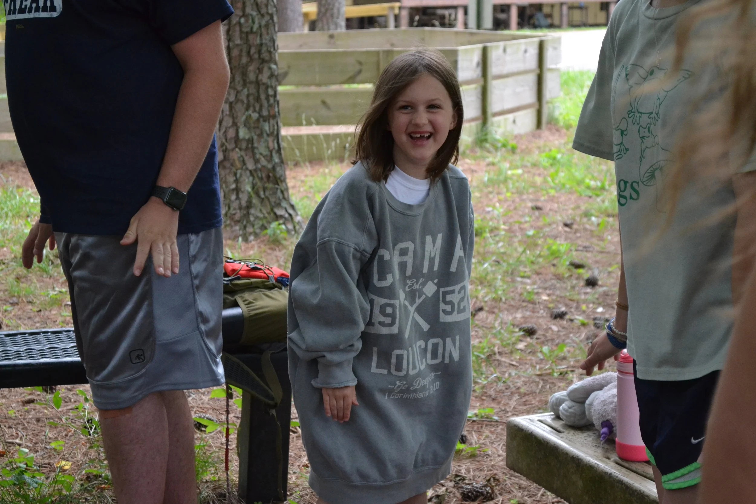 Young girl smiling and wearing a gray sweatshirt with 'CAMA 1952 LOICON' printed on it, standing outdoors among other people in a wooded area.