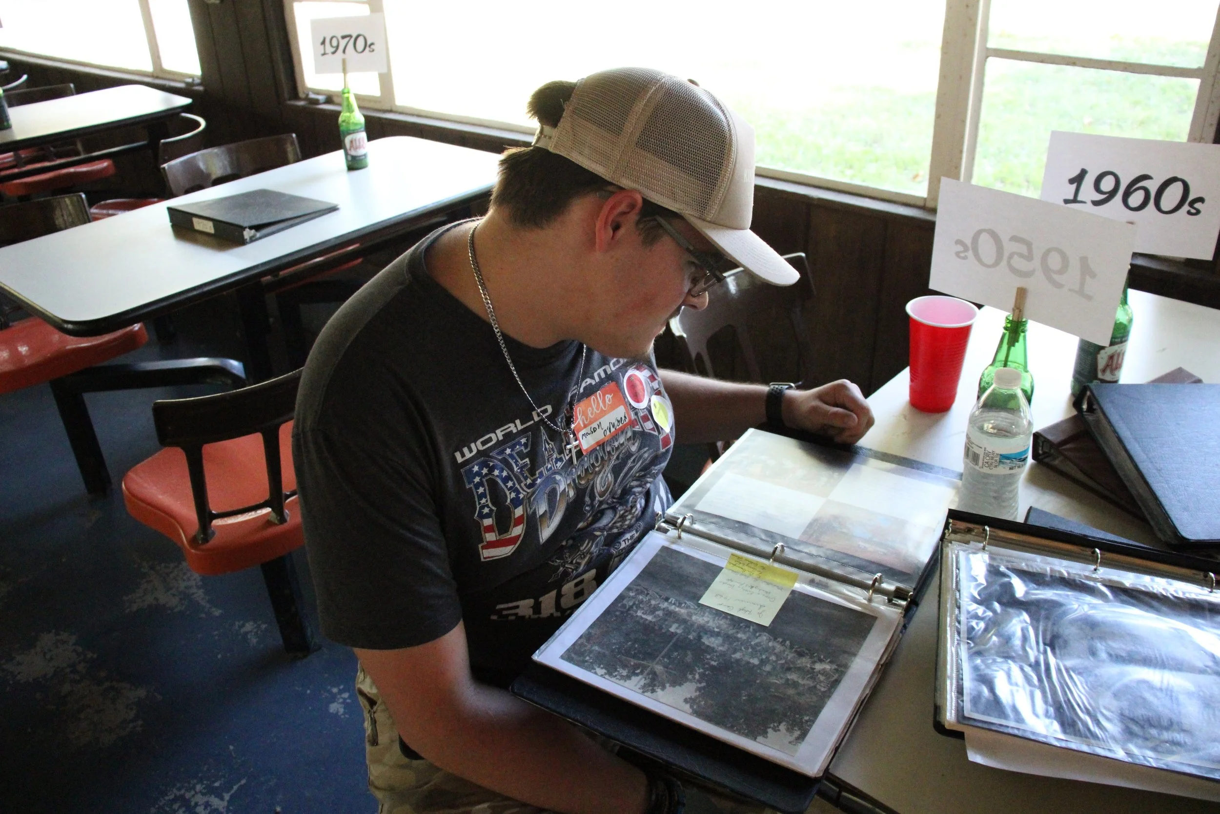 A man wearing a baseball cap, glasses, and a black T-shirt with patriotic graphics, sitting at a table and looking through photo albums. The table has water bottles, a red cup, and binders. There are signs on the table indicating different decades, including the 1970s, 1960s, and 1950s, with the 1950s sign flipped over.
