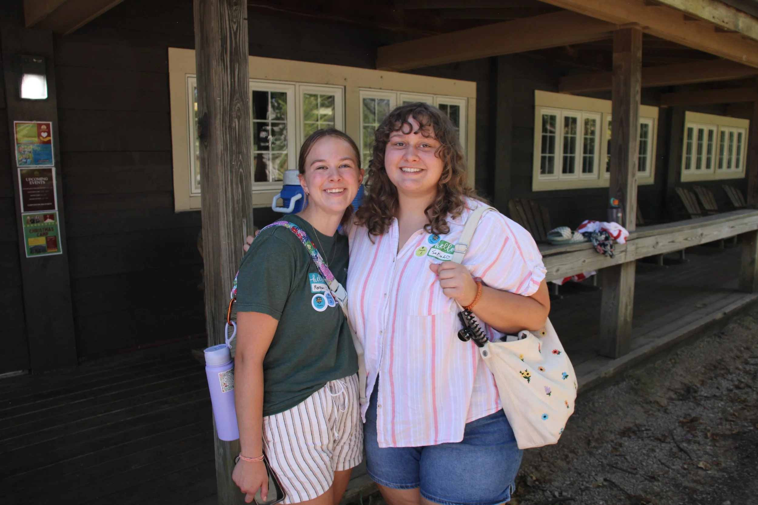 Two young women smiling and standing close together outdoors in front of a wooden building with large windows.