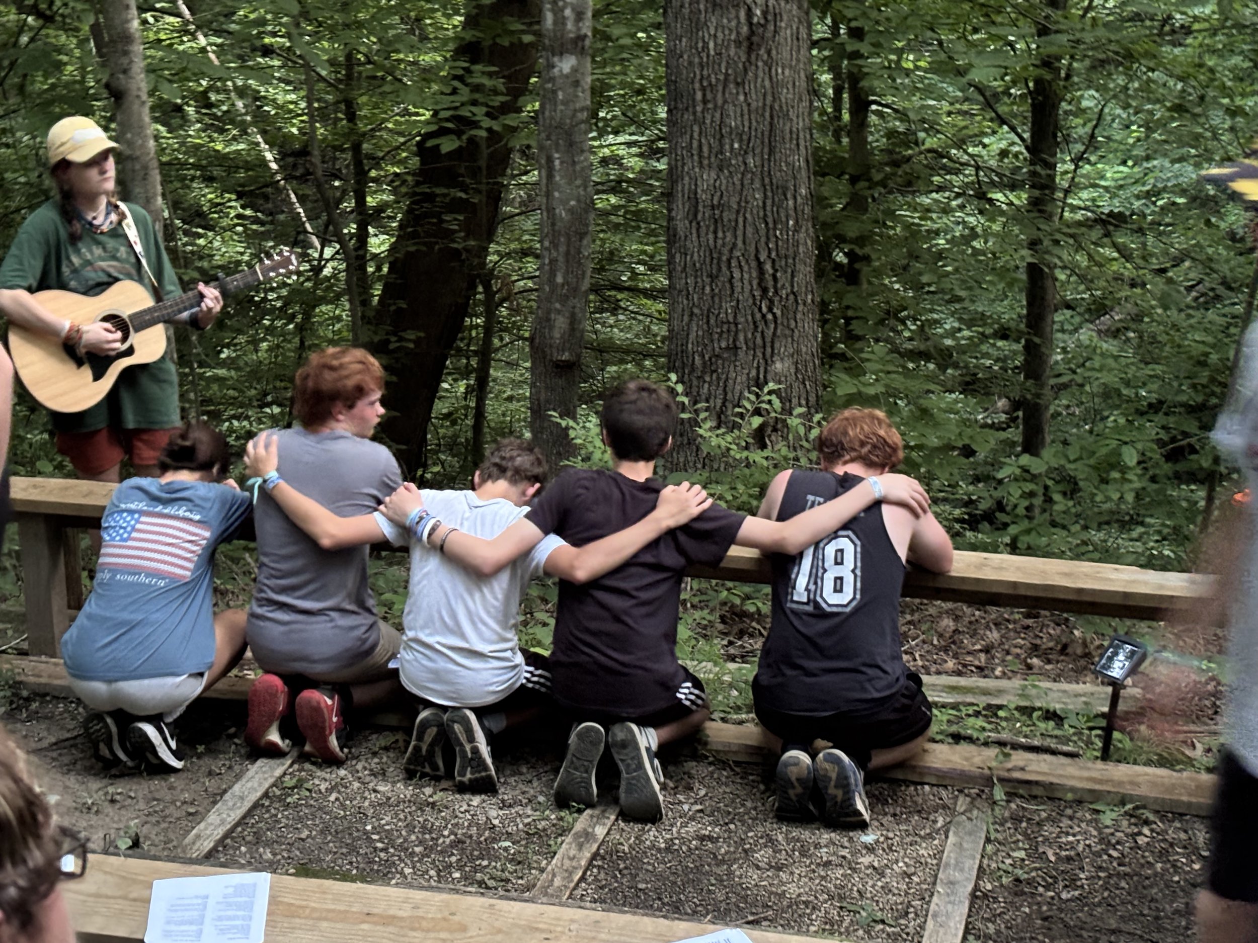 A group of children kneeling with arms around each other's shoulders, watching a person playing guitar in a wooded outdoor setting.