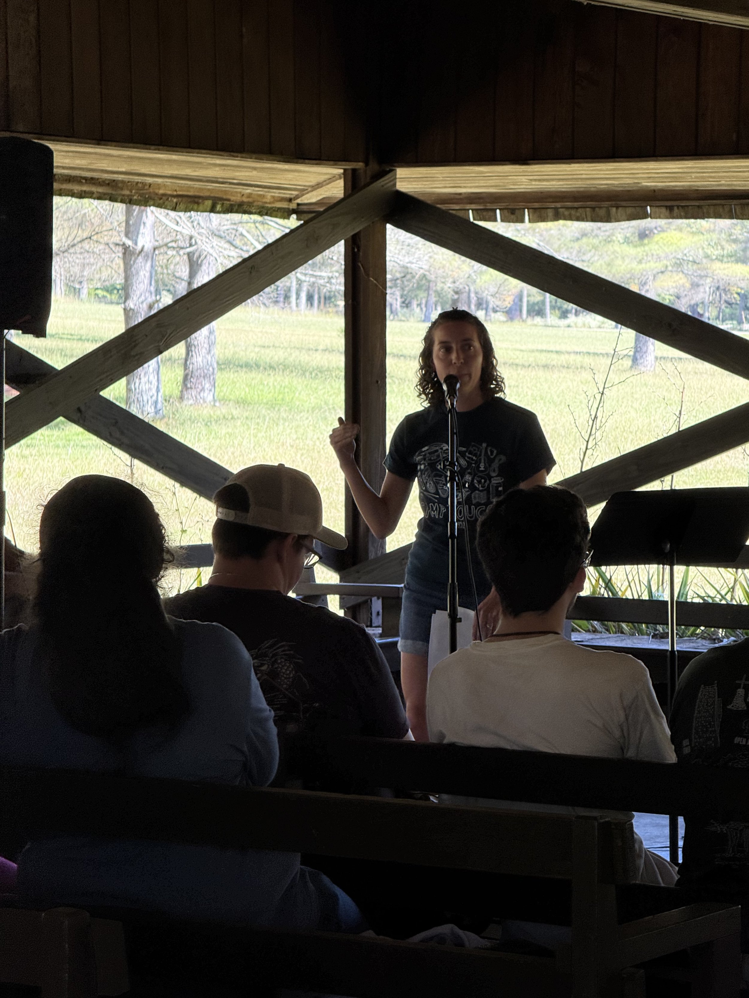 A young woman with curly hair speaking into a microphone on a small stage or platform, with a scenic outdoor background visible through the open sides of the structure. Several audience members are seated and listening to her.